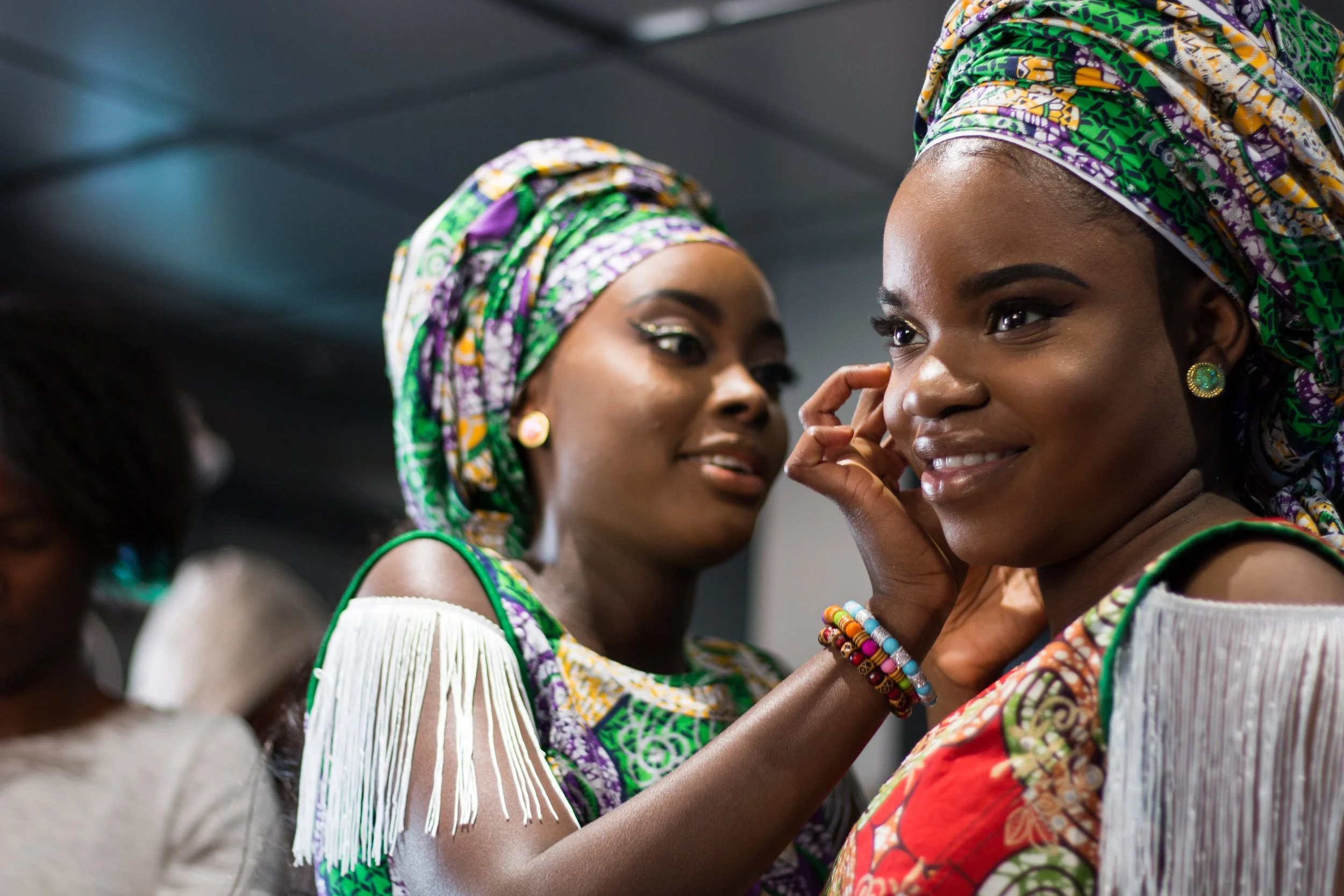 Two women wearing traditional African head wraps and clothing, one arranging the other's earring, smiling and engaging nearby others.