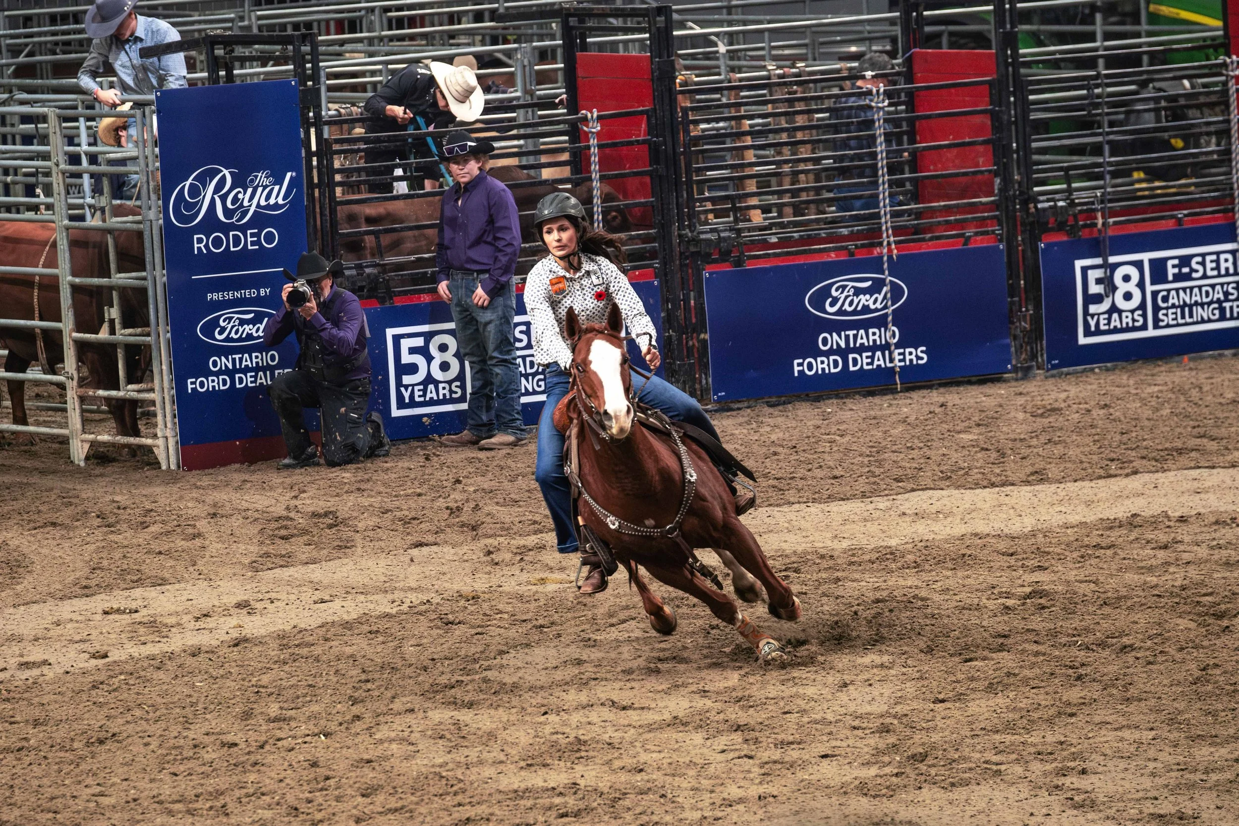 A female horseback rider wearing a helmet, white patterned shirt, and jeans riding a brown horse at a rodeo event. In the background, there are spectators, photographers, and banners advertising The Royal Rodeo and Ontario Ford Dealers.