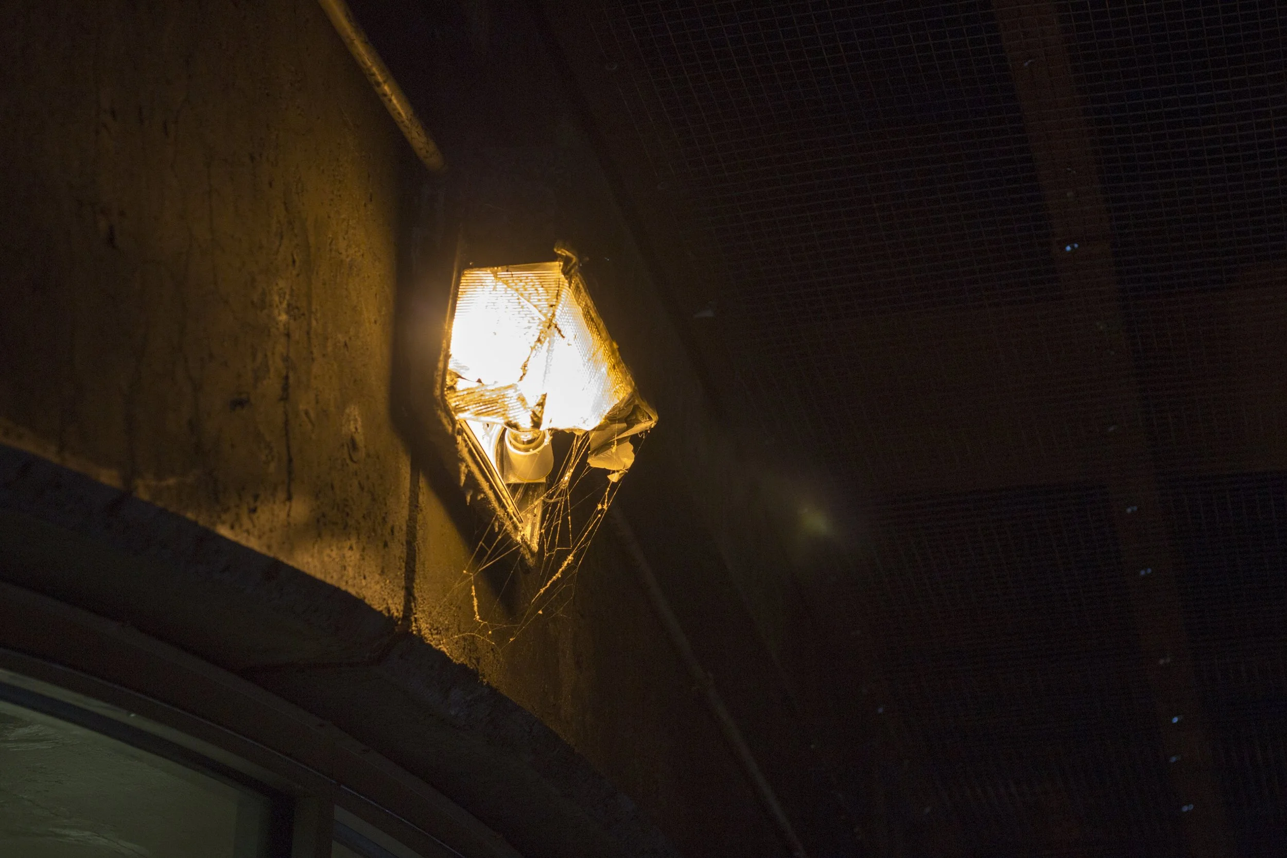 A damaged outdoor wall-mounted light fixture with a cracked glass cover emitting bright light, surrounded by cobwebs, attached to a textured wall underneath a dark wooden ceiling.