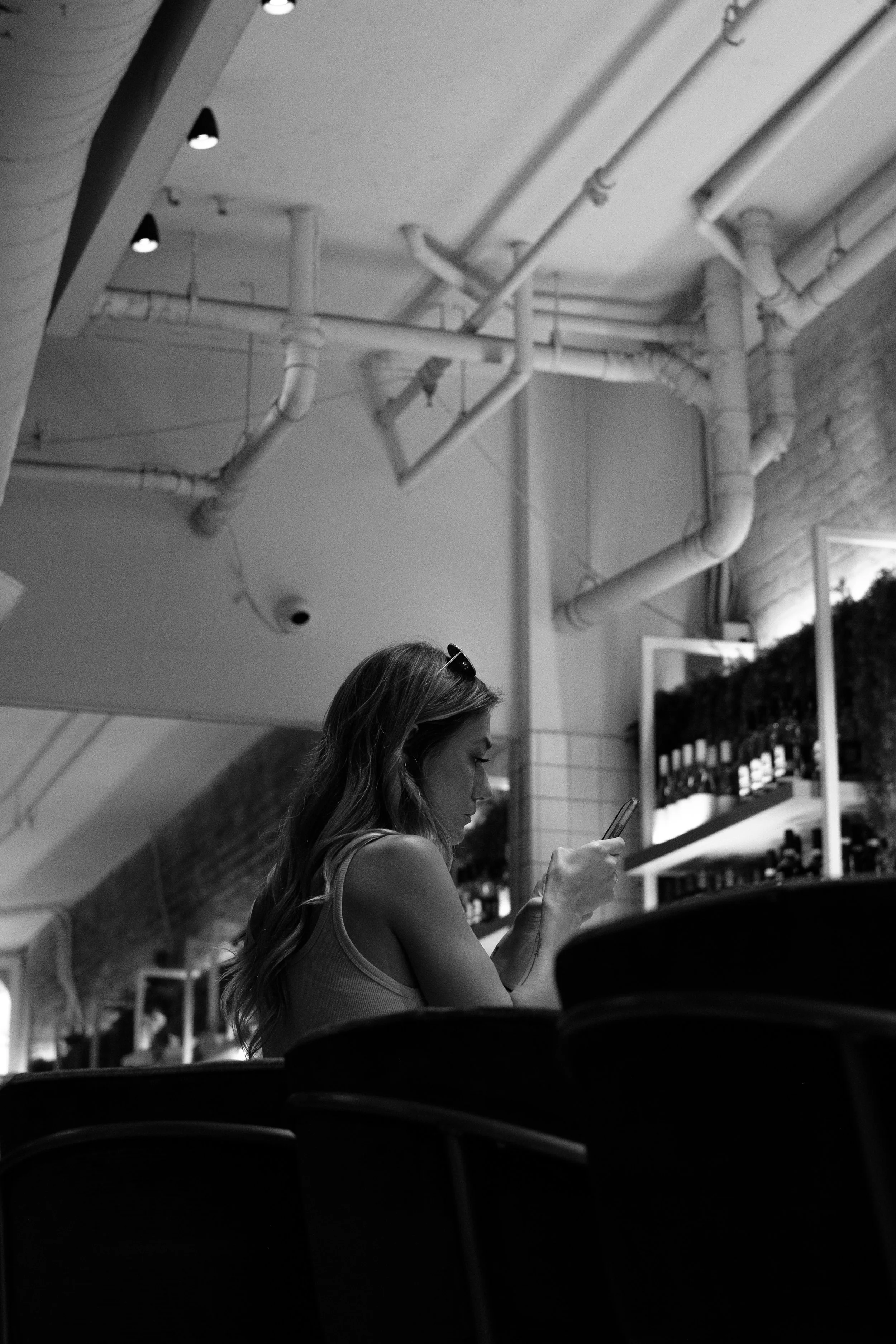 A woman with long hair, wearing sunglasses on her head, sitting at a table in a modern indoor setting with exposed pipes on the ceiling, looking at her phone.