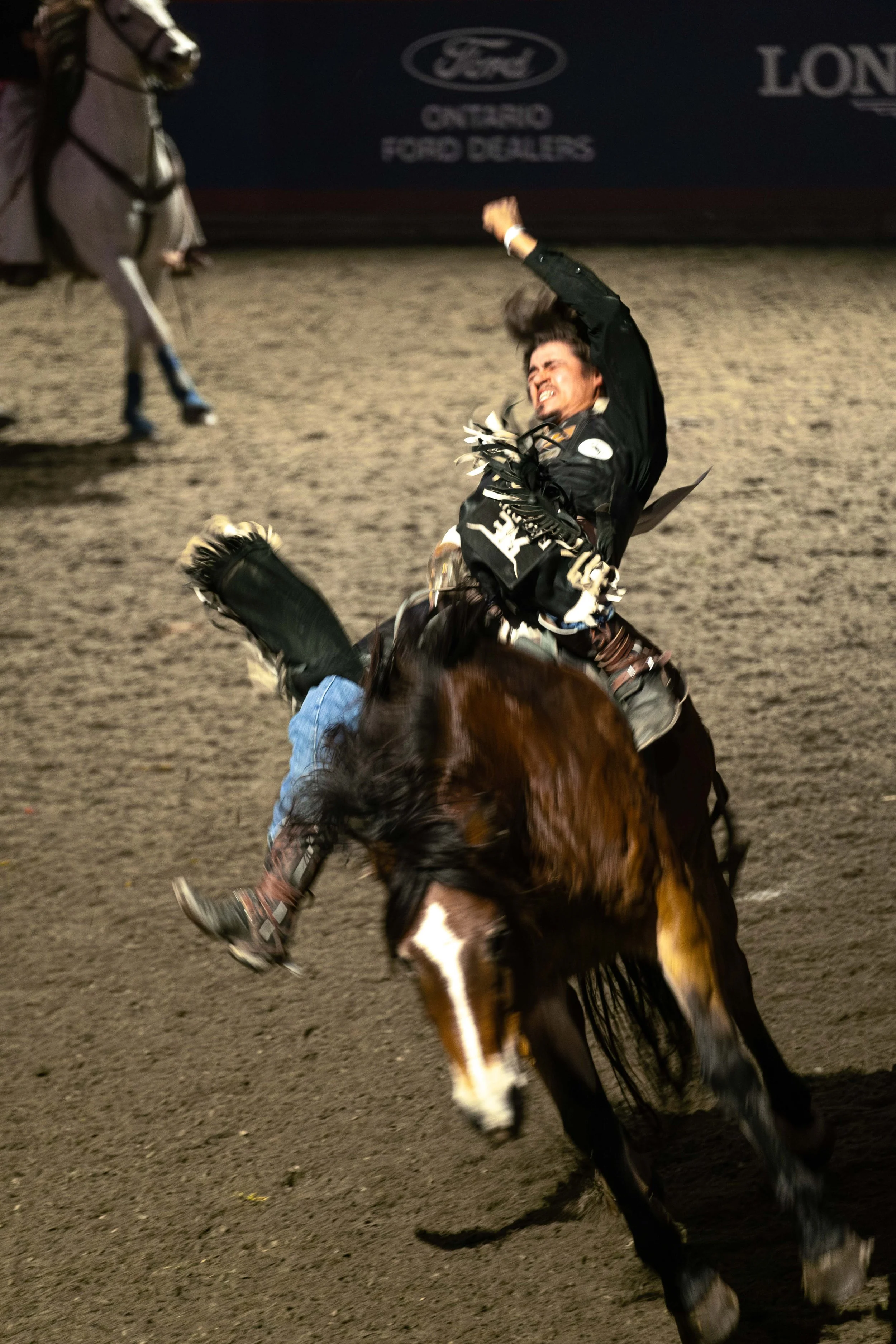 A rodeo rider in black attire falling off a brown and white horse on a dirt arena, with a sponsor sign in the background.
