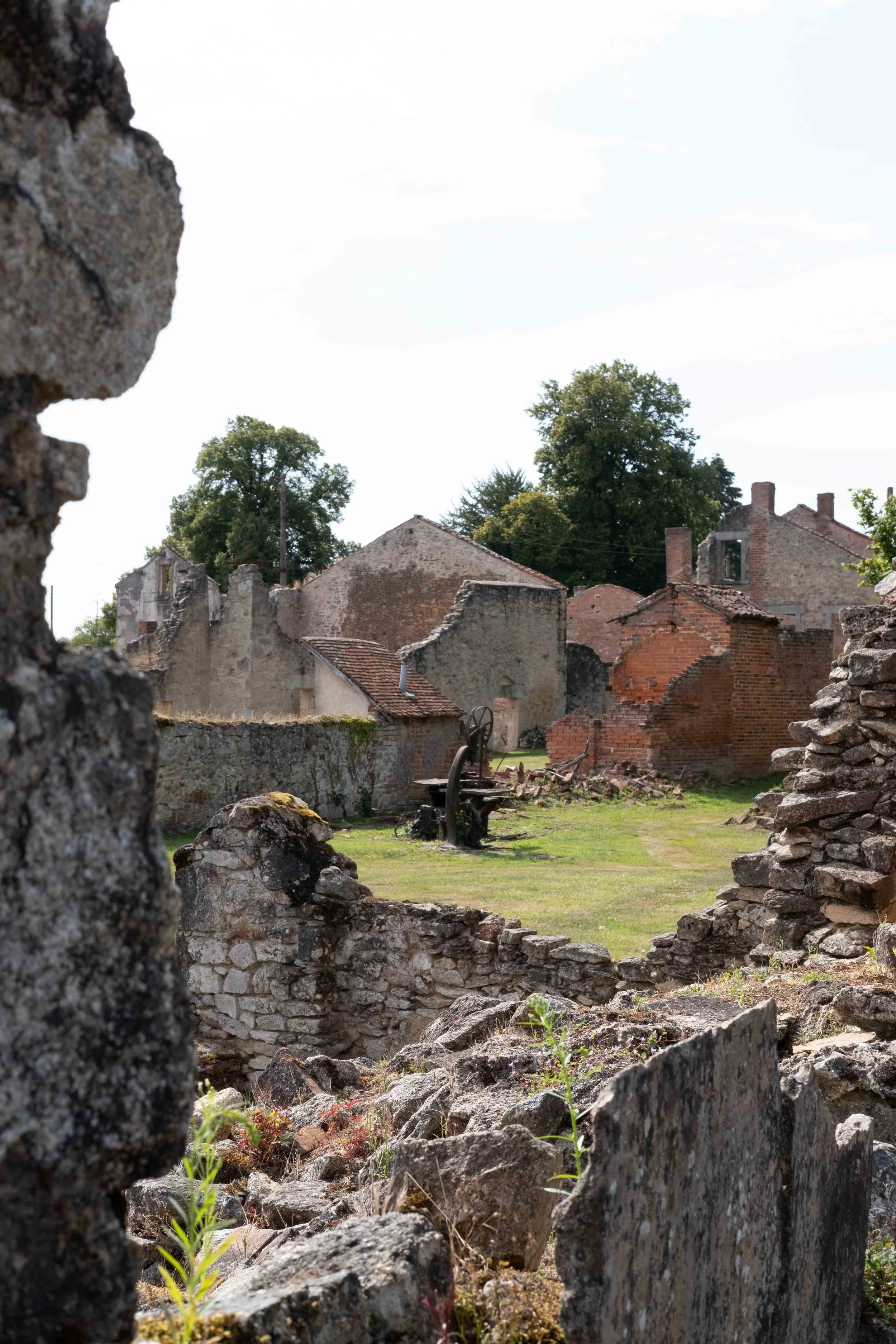 A view of ruins with weathered stones and walls, an old mechanical device in the open area, and trees in the background.