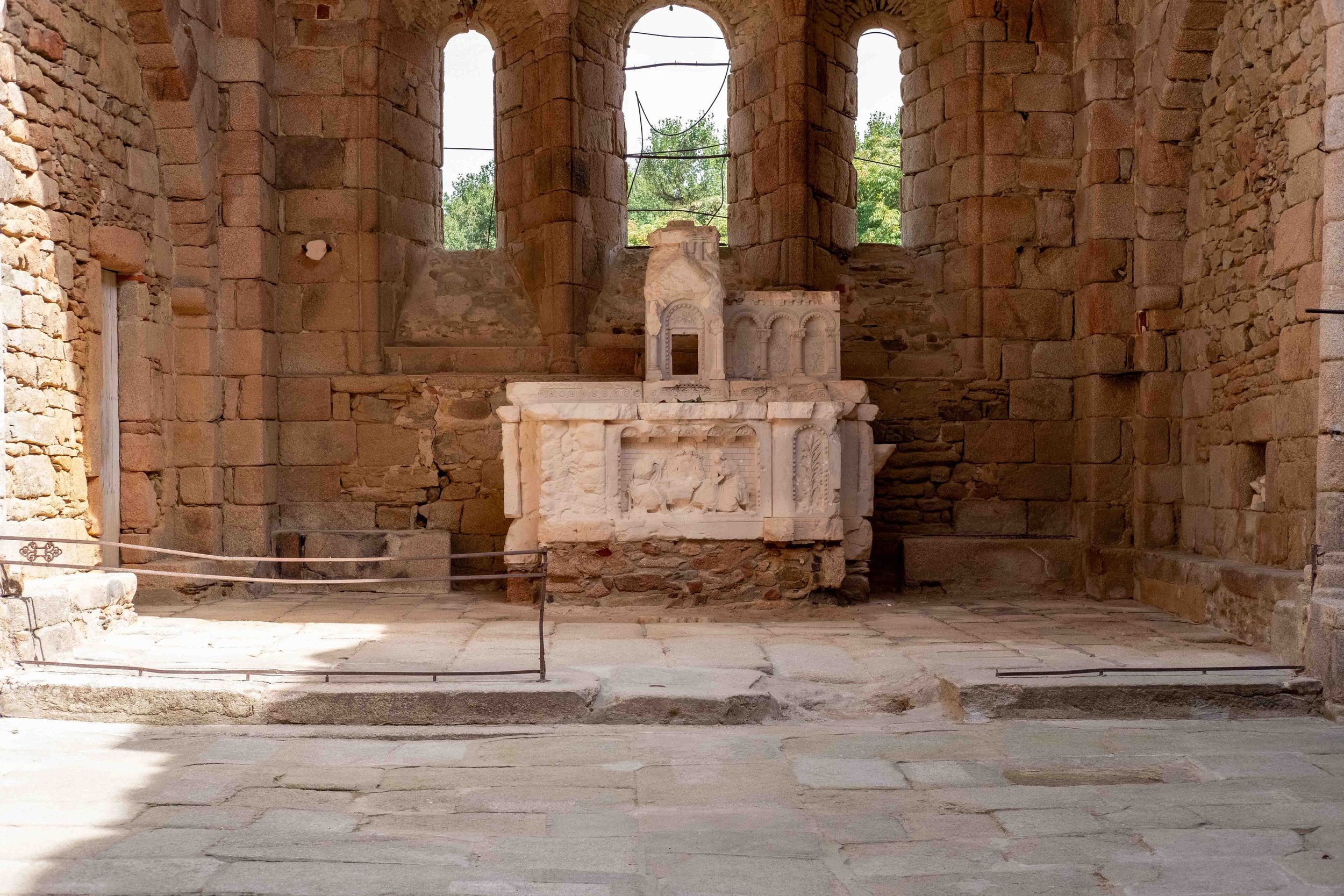 Ancient stone temple ruin with a white marble altar in the center, featuring intricate carvings of animals and decorative arches, set inside a structure with tall, arched windows and stone walls.