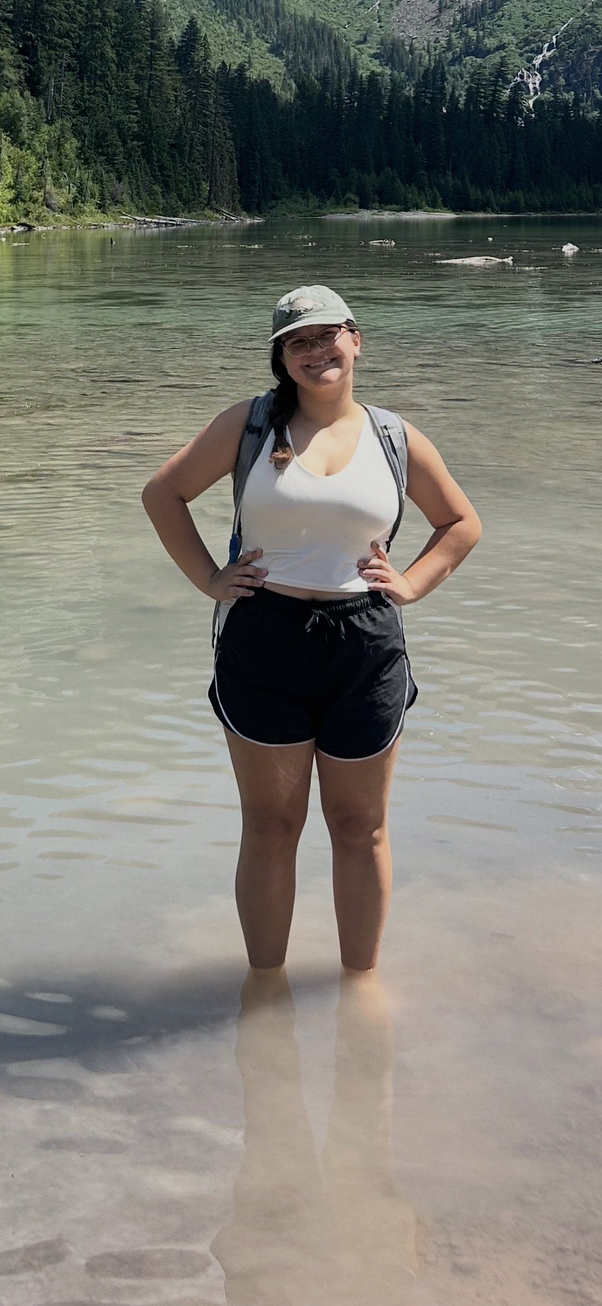 A woman standing in shallow water by a lake with a forest and waterfall in the background. She is smiling, wearing glasses, a light-colored cap, white tank top, and black shorts, with a backpack.