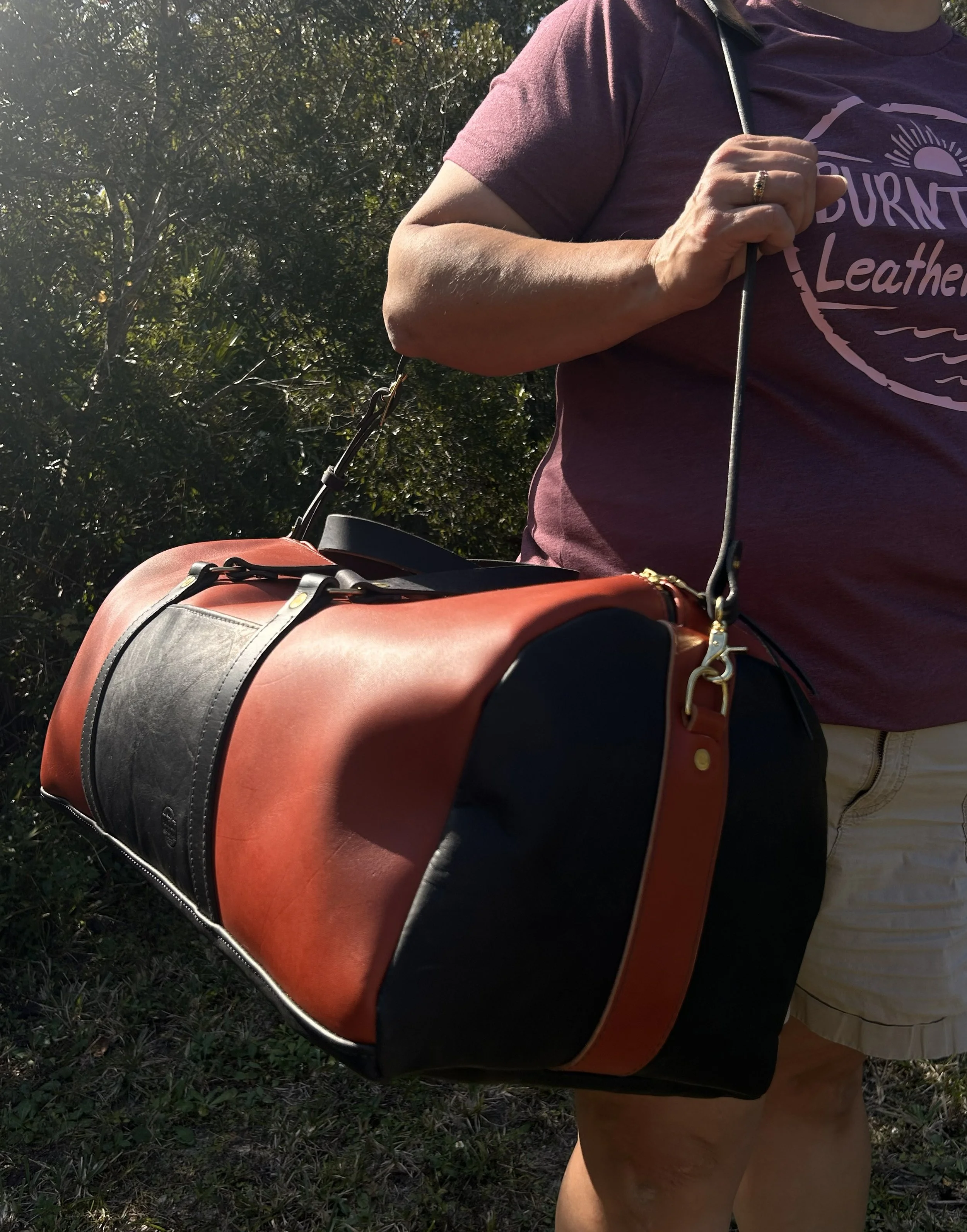 Person walking outdoors carrying a large orange and black leather duffel bag on their shoulder, wearing a maroon t-shirt and beige shorts, with trees and sunlight in the background.