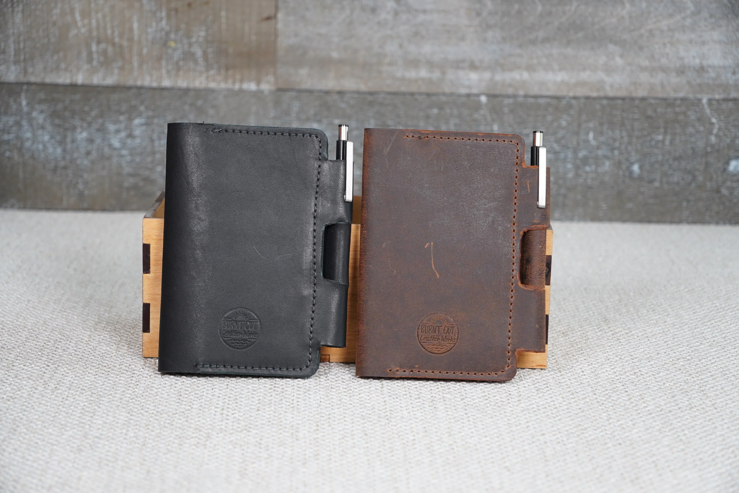 Two leather field notebook covers, one black and one brown, placed on a wooden stand against a background of a gray brick wall.