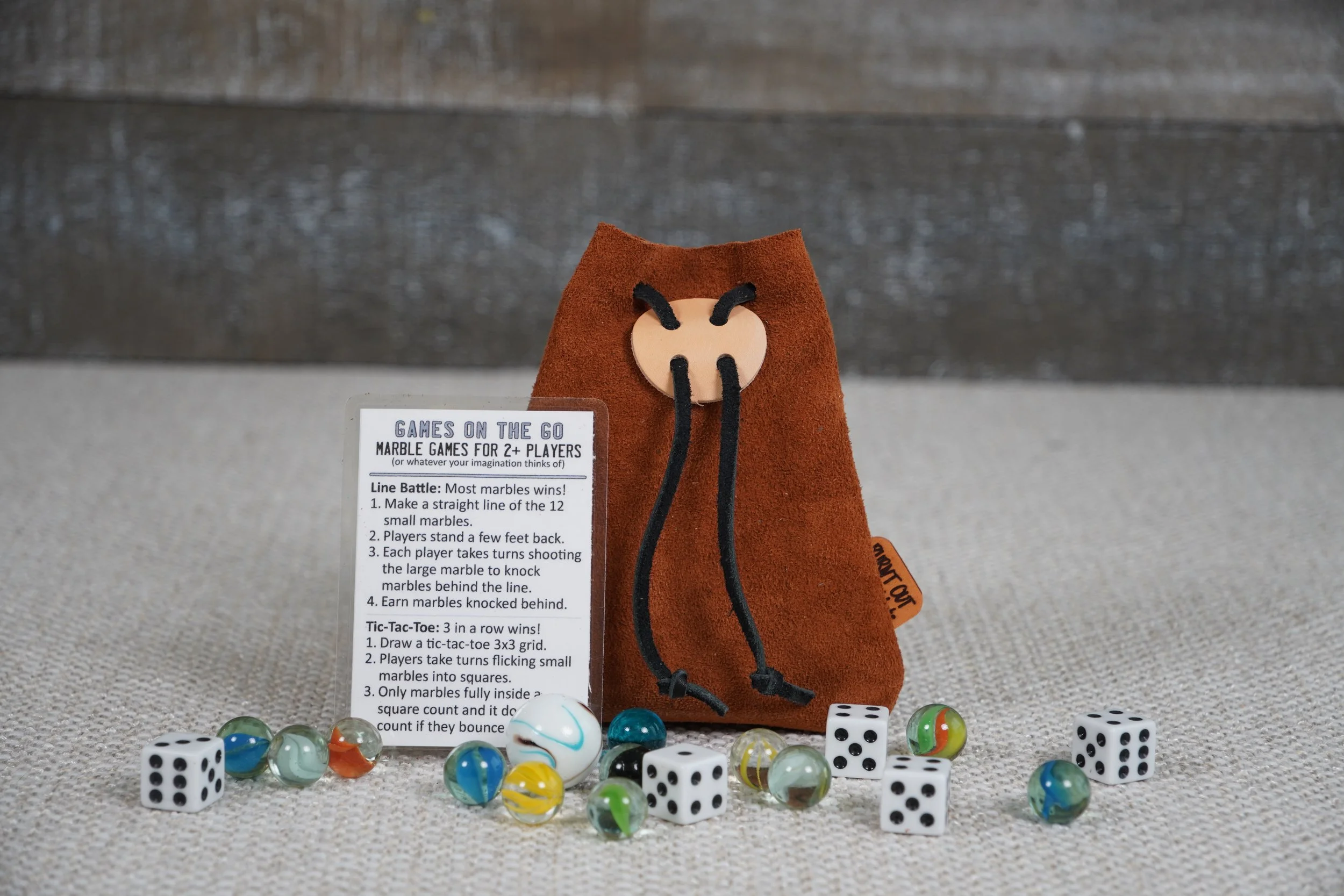 A plush brown velvet bag with a wooden ant face and black string antennae, surrounded by various game pieces, including dice and marbles, and a game instructions sheet for marble-based games.