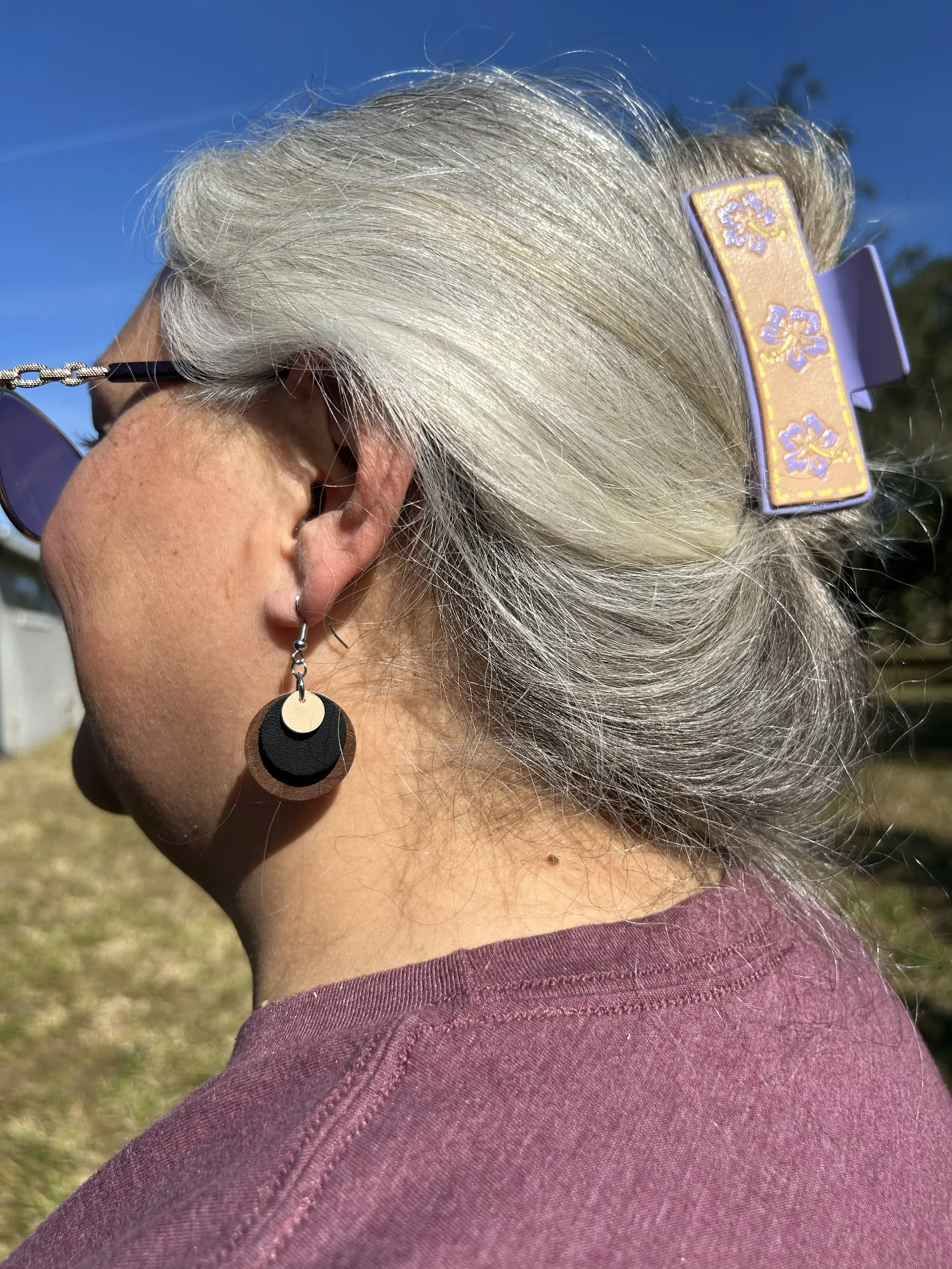 Close-up of a person with silver hair in a bun, wearing sunglasses, a decorative hair clip with purple and yellow flowers, and dangling earrings, outdoors on a sunny day.
