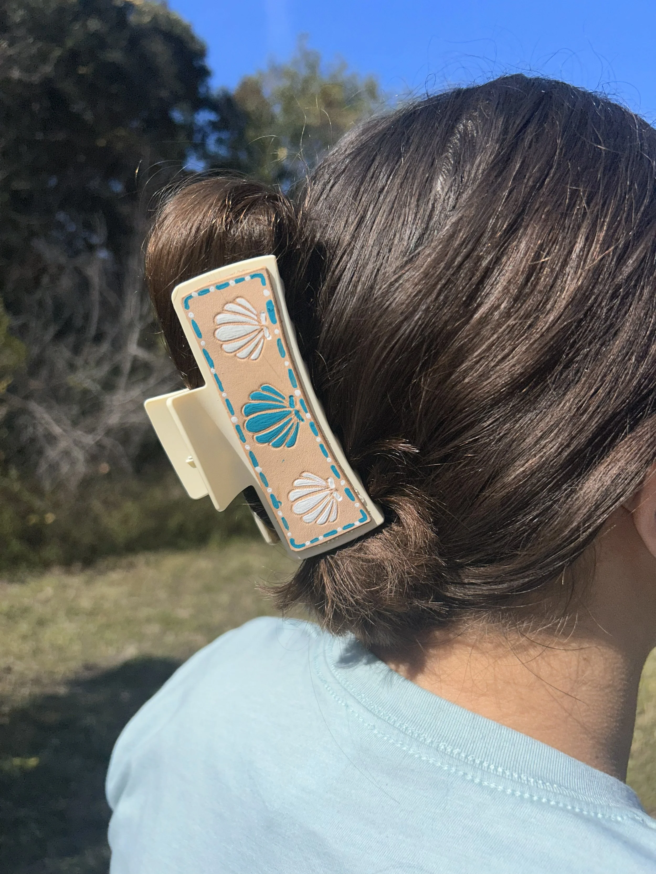 Close-up of a person's brown hair held by a decorative beige hair clip with blue and white shell designs, outdoors with blue sky and trees in the background.