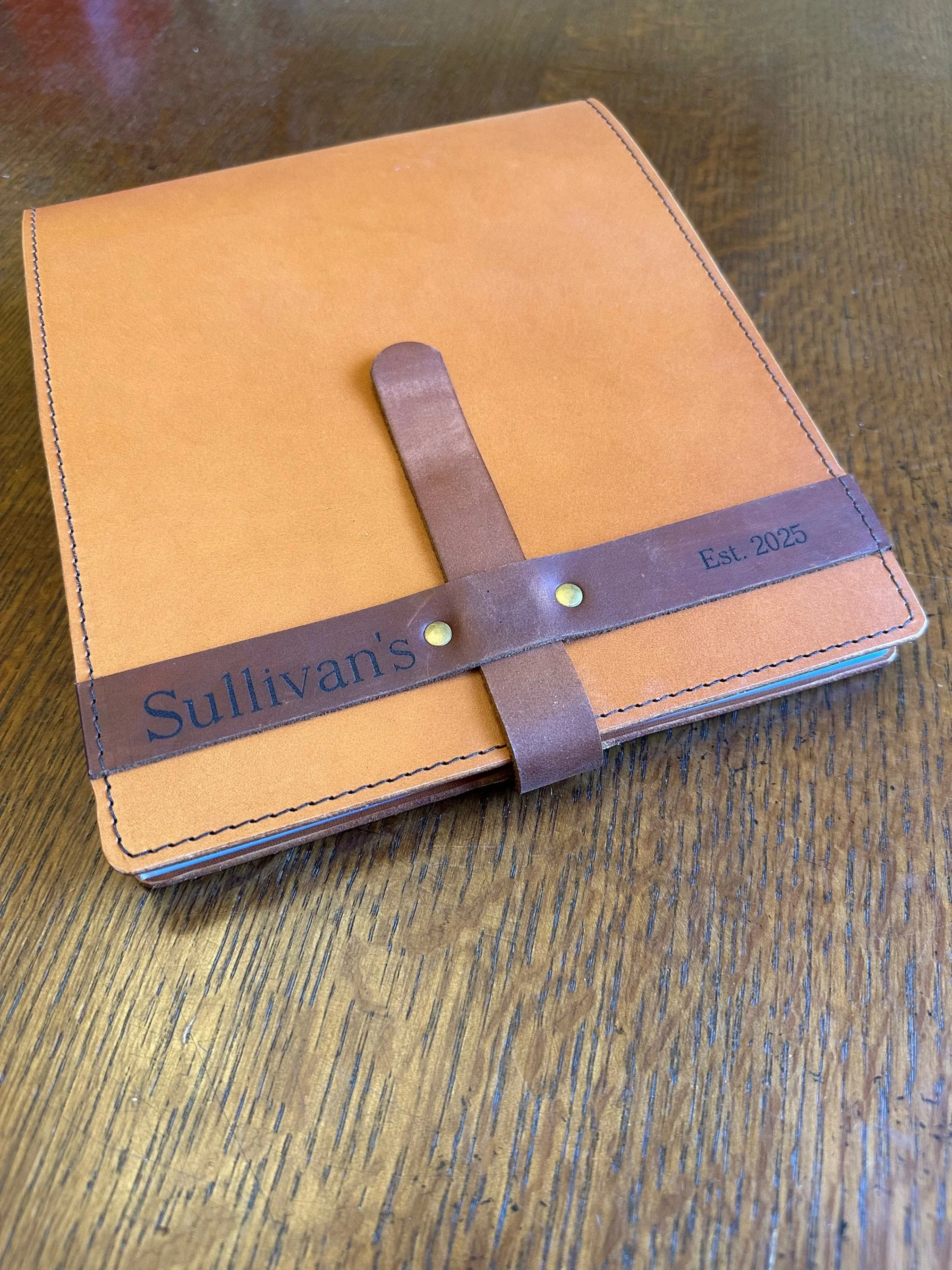 A tan-colored leather-bound notebook with a brown leather strap and brass rivets, labeled "Sullivan's Est. 2025," resting on a wooden surface.