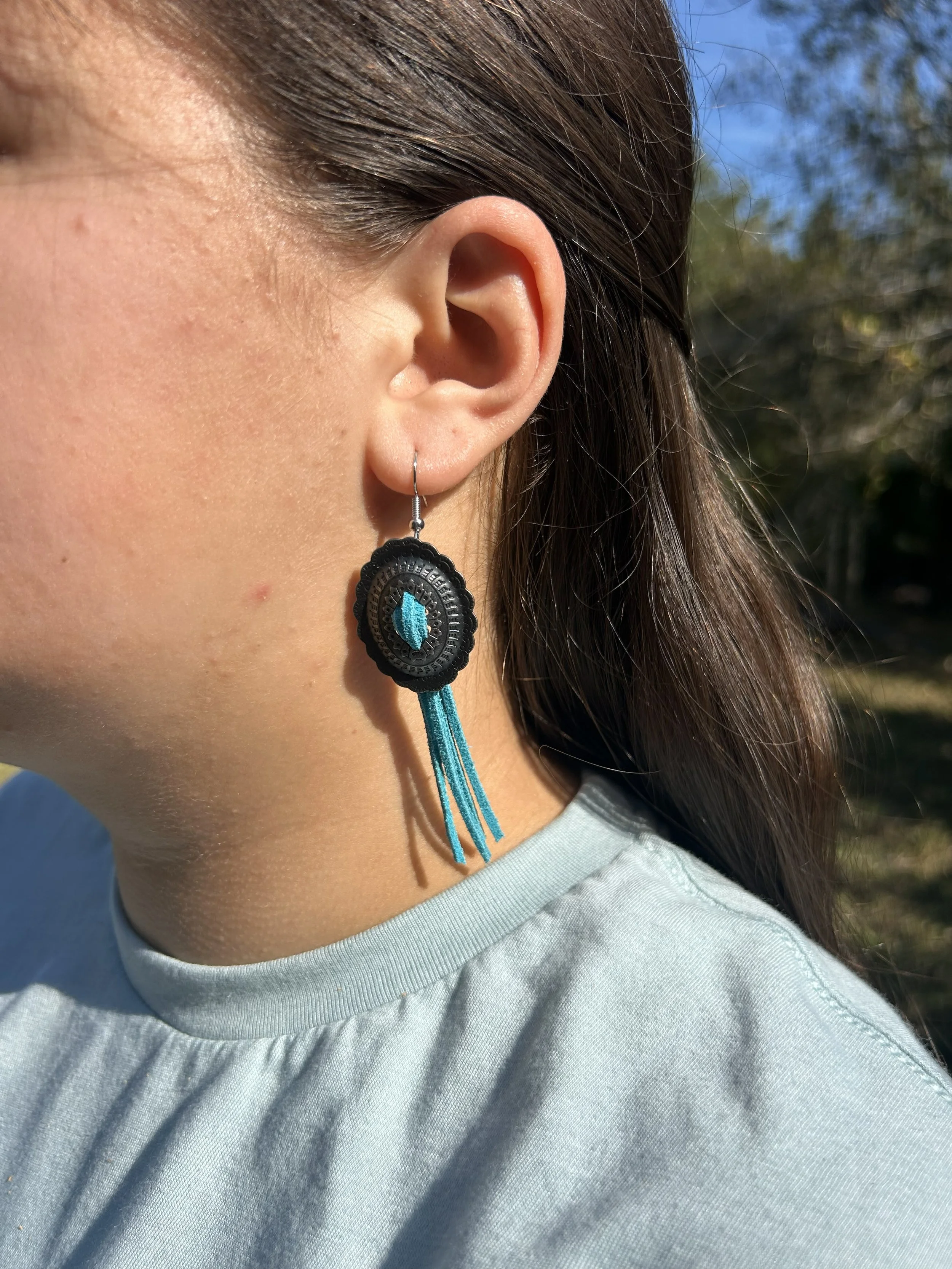 Close-up of a woman's ear with an earring that has a black and blue design and hanging blue threads, outdoors in sunlight.