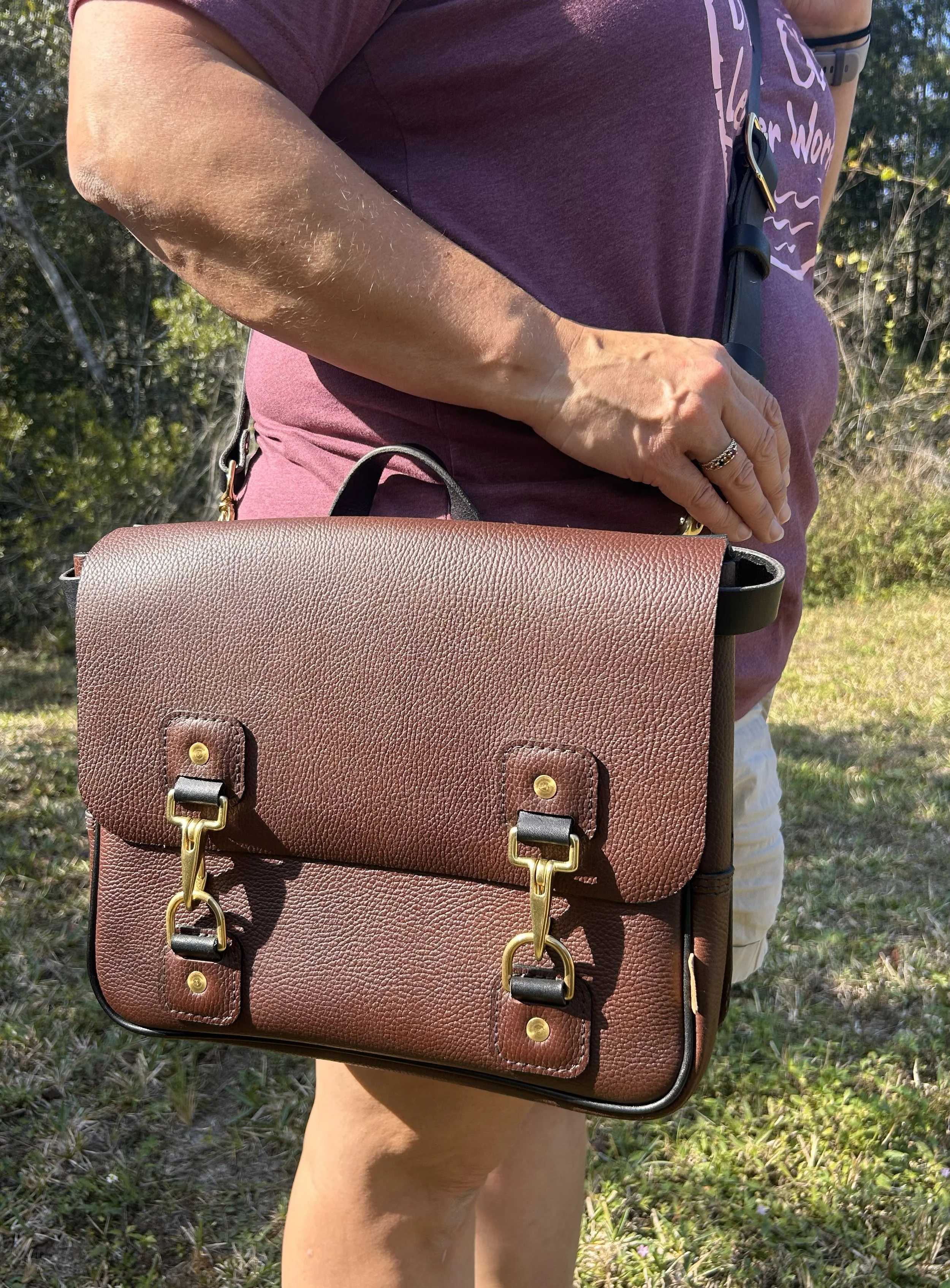Close-up of a person holding a brown leather handbag with gold hardware, outdoors in a grassy area with trees in the background. The person is wearing a purple shirt and a ring on their left hand.