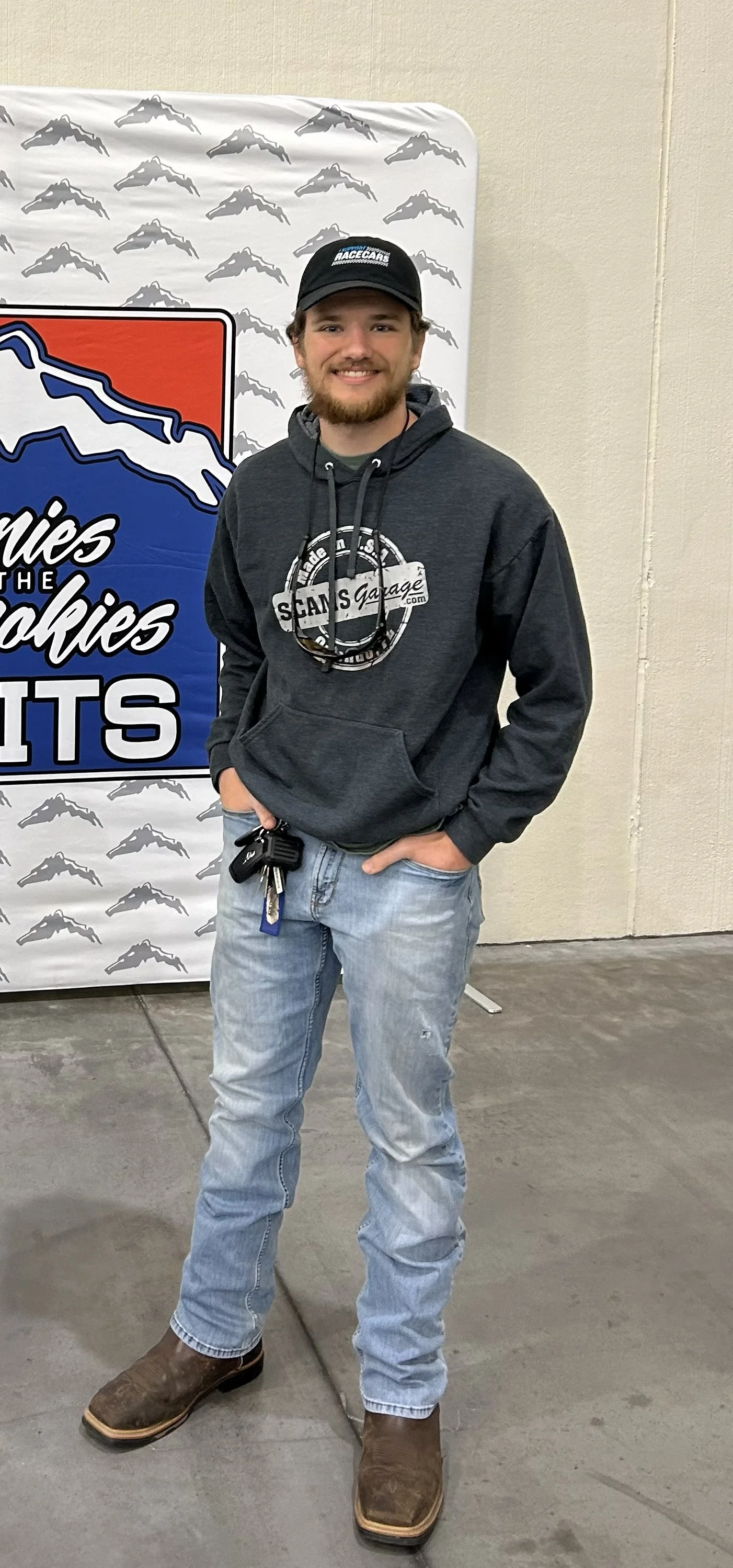 A young man with a beard and light brown hair posing indoors in front of a sign that says 'Scanies of the Rockies'. He is wearing a black cap, a gray hoodie with a logo that says 'Scams Garage', light-washed jeans, and brown cowboy boots. He has his hands in his pockets and is smiling at the camera.
