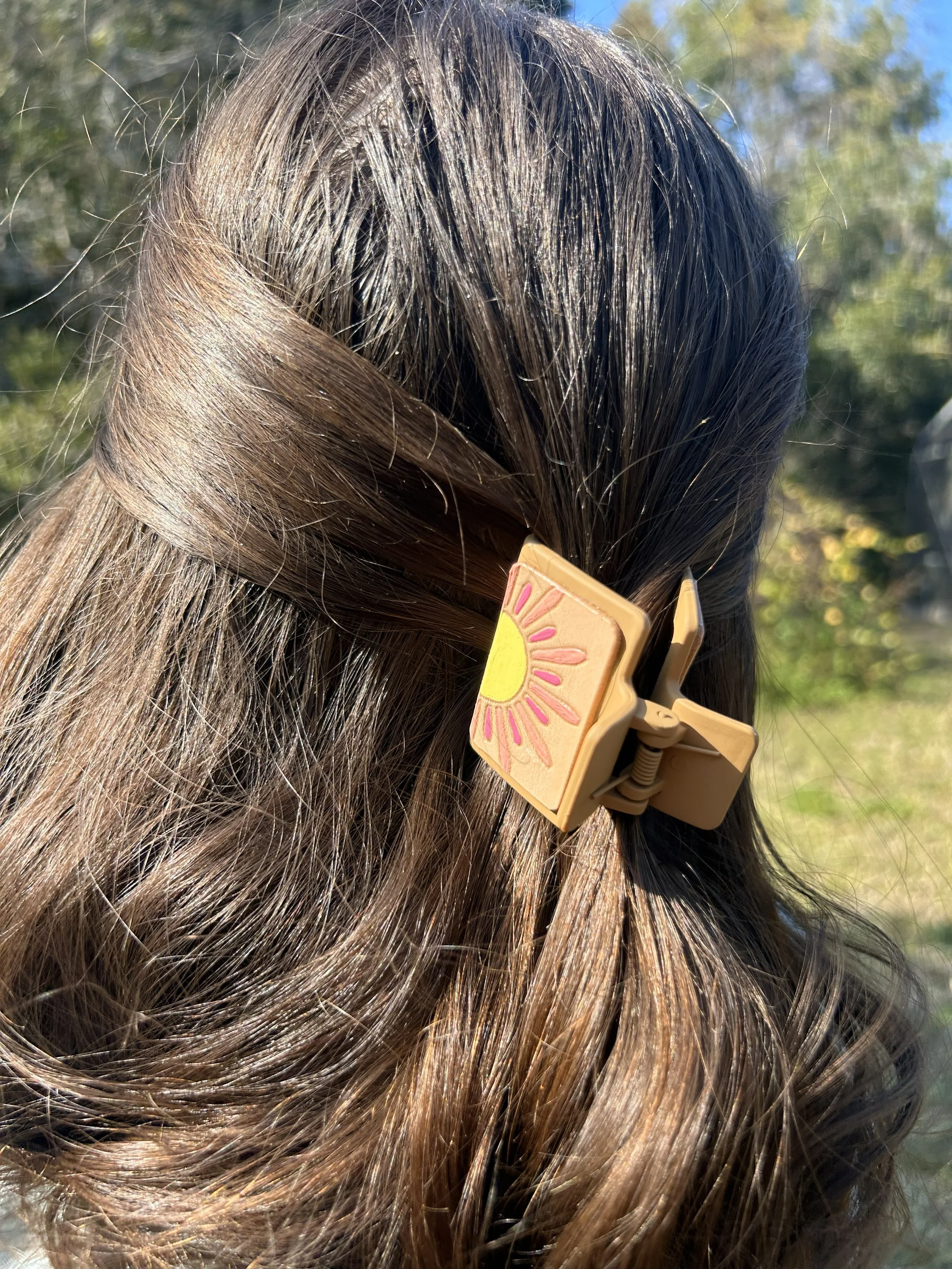 A person's brown hair secured with a beige hair clip with a painted yellow sun and pink rays design, outdoors with blurred greenery in the background.