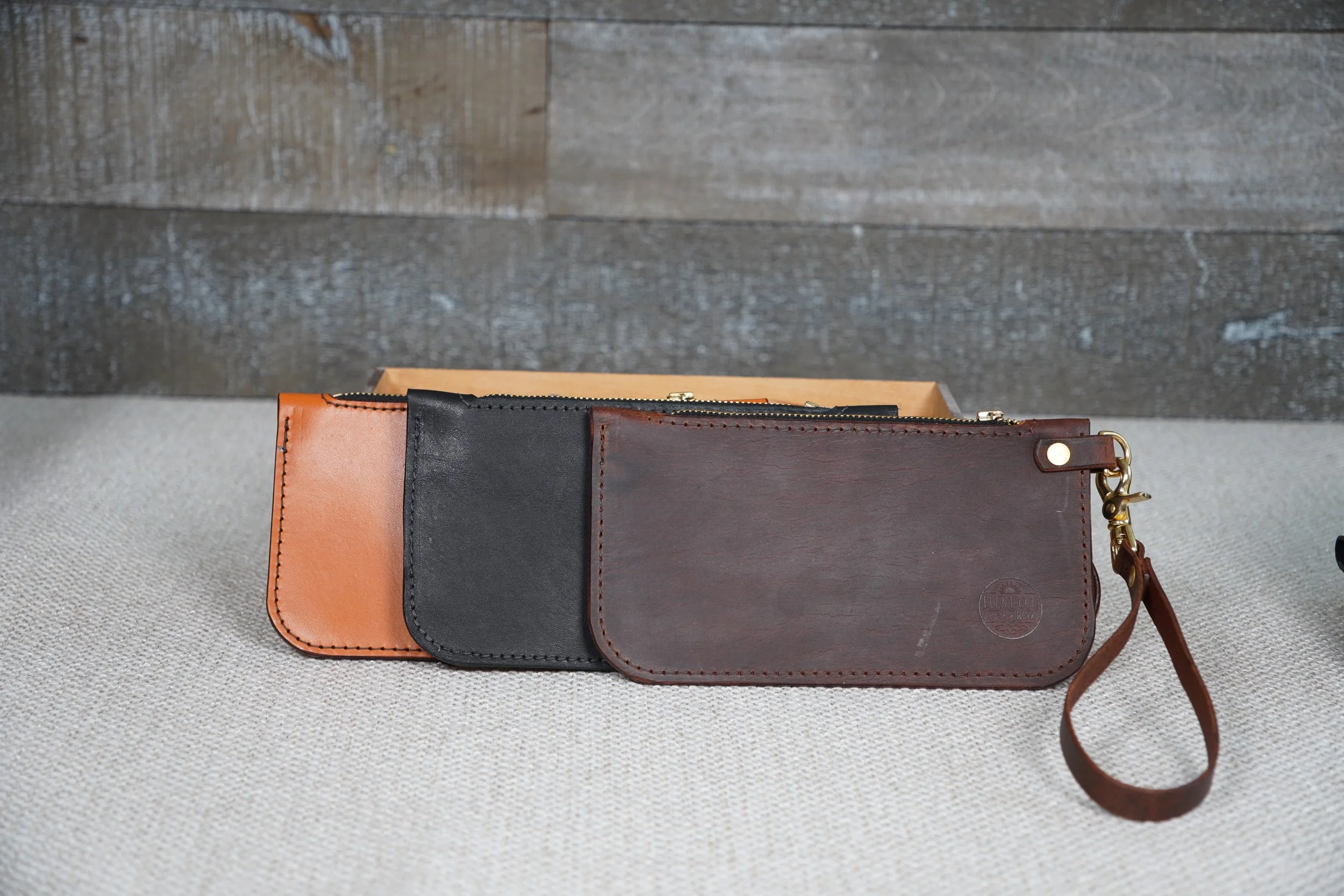 Three leather wallets in different colors, tan, black, and dark brown, with the brown wallet featuring a small circular embossed logo and a attached leather wrist strap, placed on a beige fabric surface with a wooden wall background.