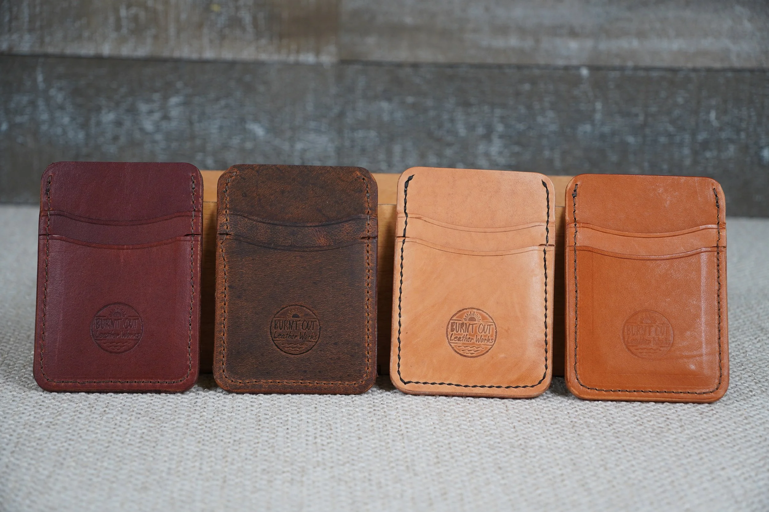 Four leather cardholders in different colors arranged in a row on a fabric surface with a wooden background