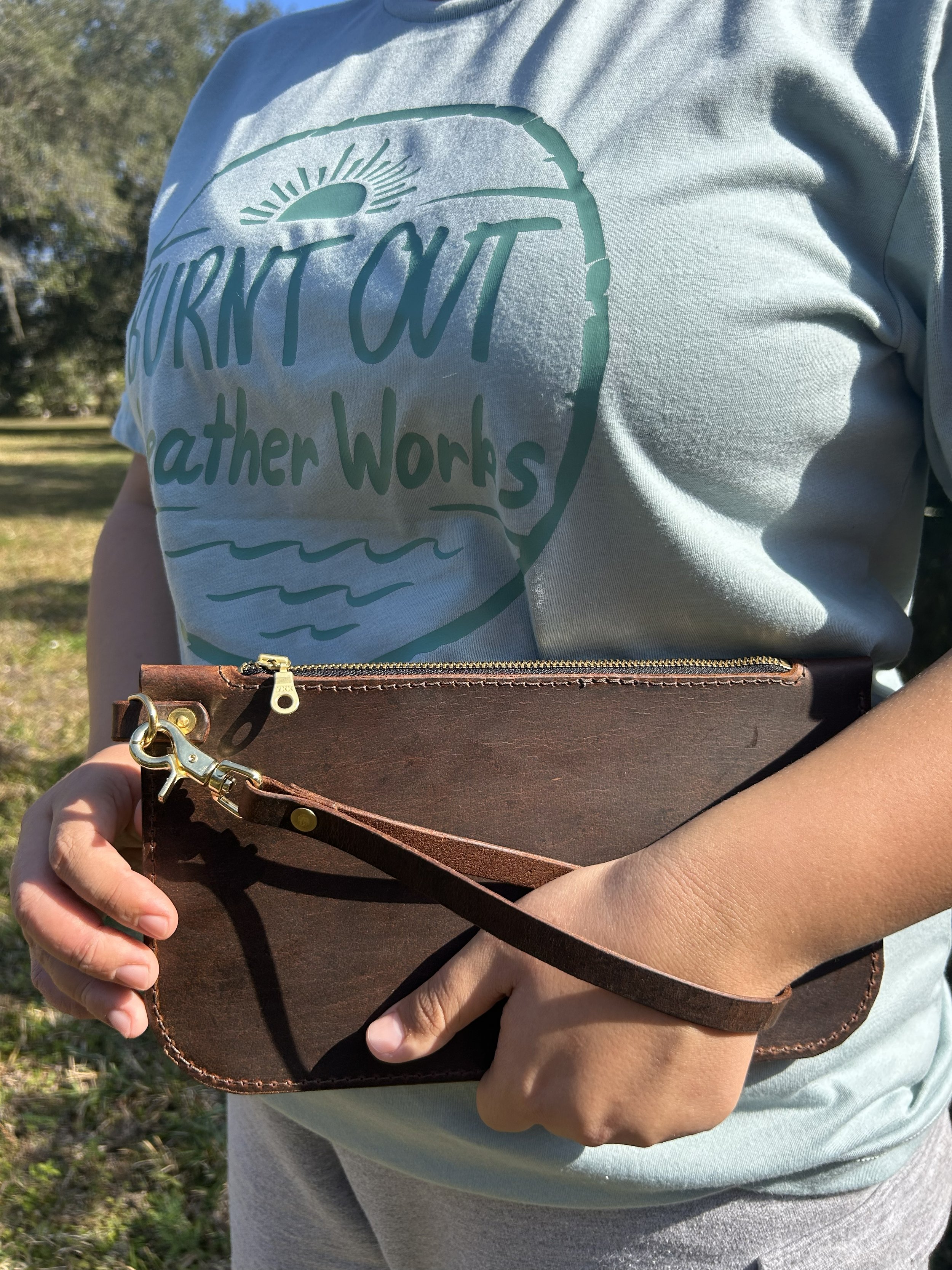 Person wearing a light blue t-shirt with a slogan and holding a brown zippered bag outdoors.