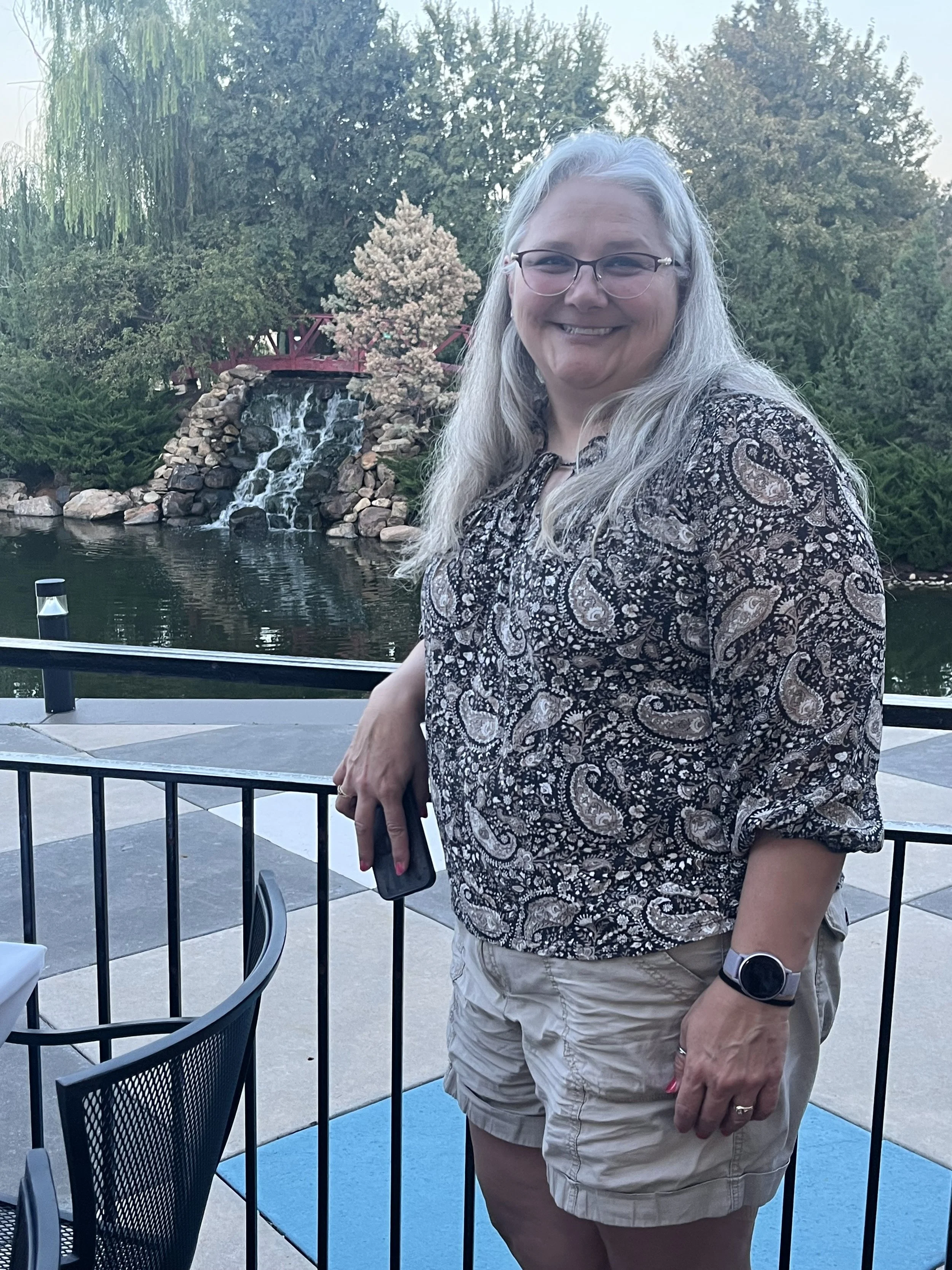 A woman with long gray hair and glasses smiling outdoors on a patio with a pond, small waterfall, and trees in the background.