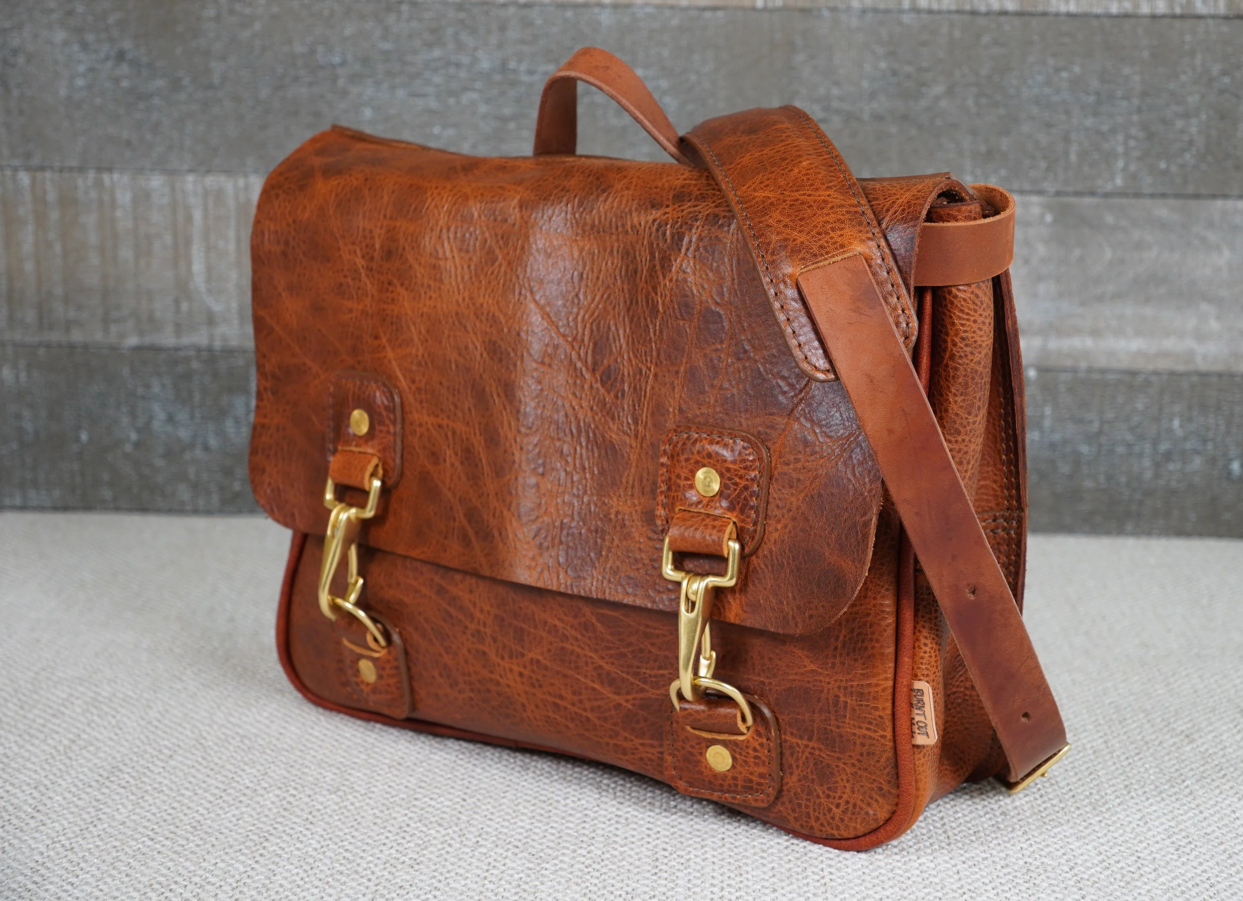 A brown leather shoulder bag with gold hardware, sitting on a textured light-colored surface.