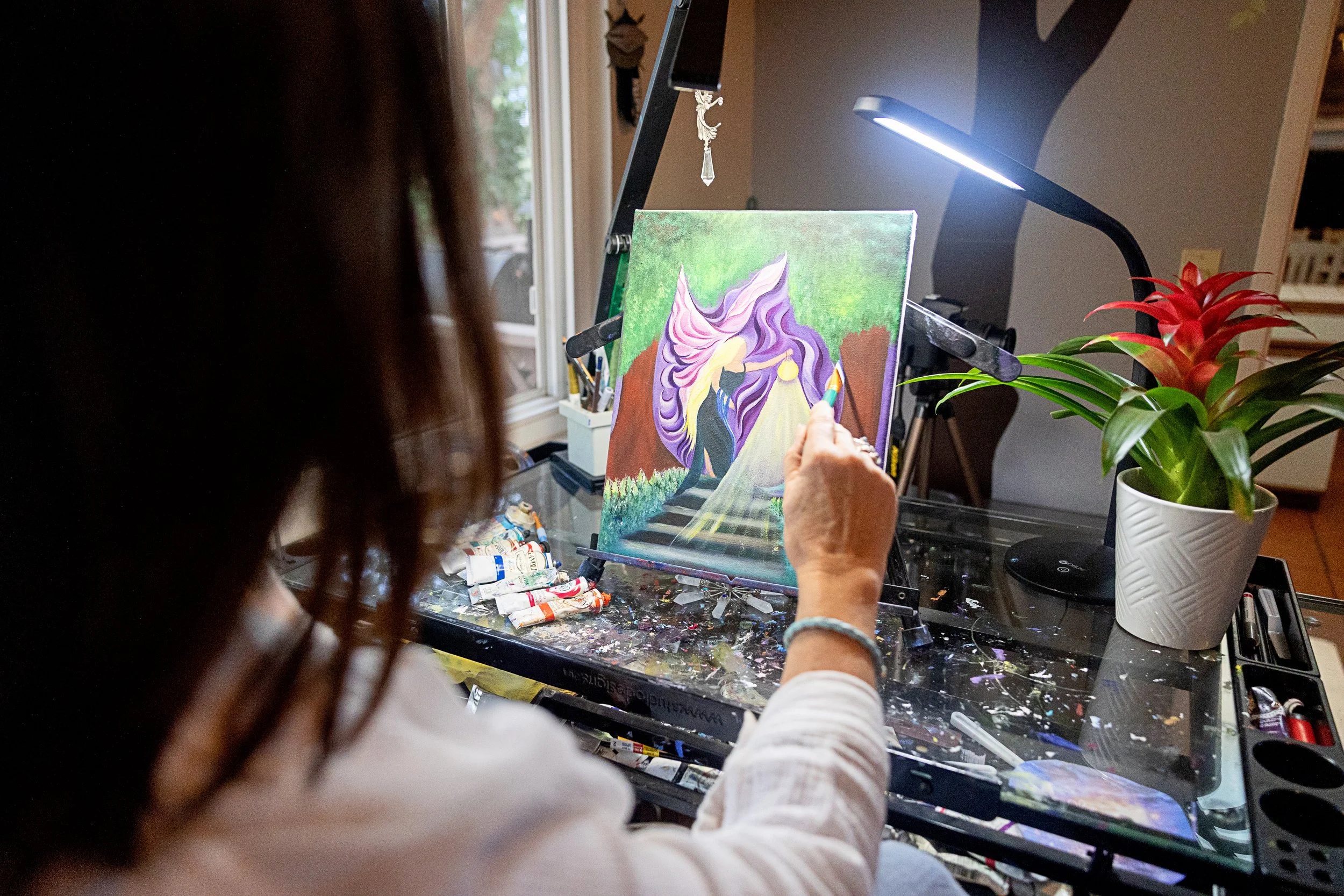 A person painting a colorful abstract portrait of a unicorn on a canvas, with art supplies and a red and green plant nearby.