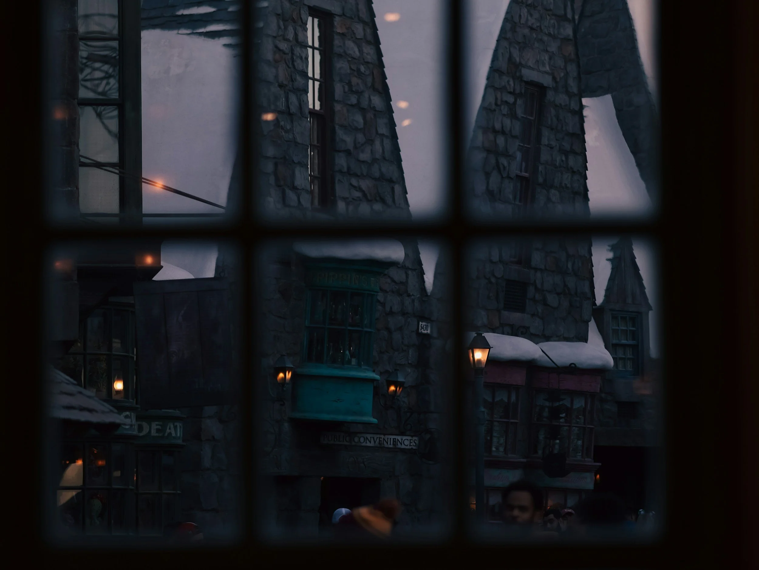 View through a grid window showing buildings with stone and wood facades, some with snow on the roofs, and lantern-style lights during dusk or early evening.