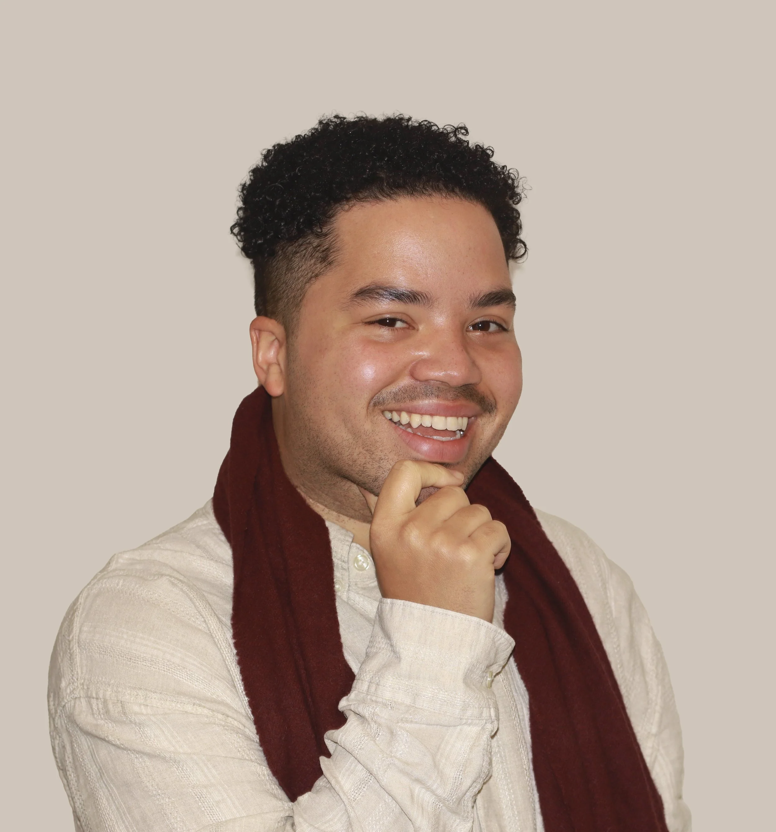 A young man smiling confidently with one hand on his chin, wearing a beige shirt and a dark red scarf, against a plain background.