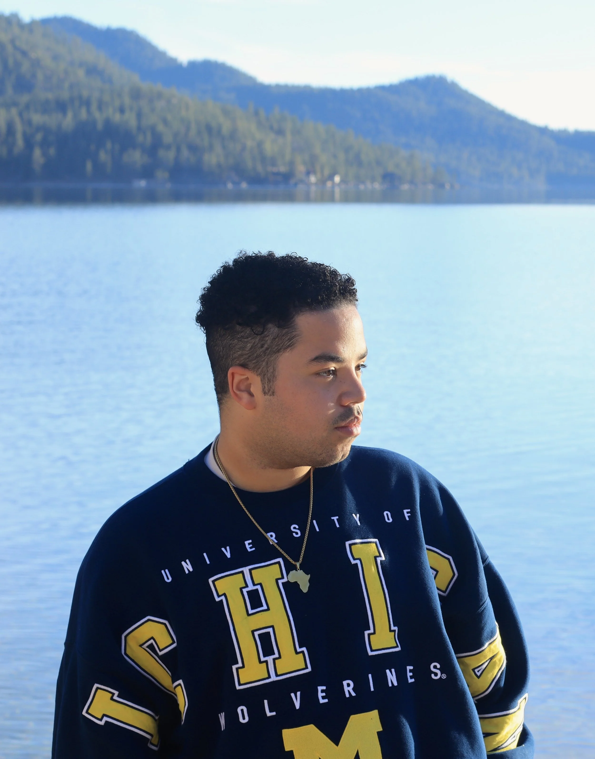 A young man with short curly hair and a necklace with a map pendant, wearing a navy blue sweatshirt with yellow and white text, standing in front of a large lake with mountains in the background on a clear day.