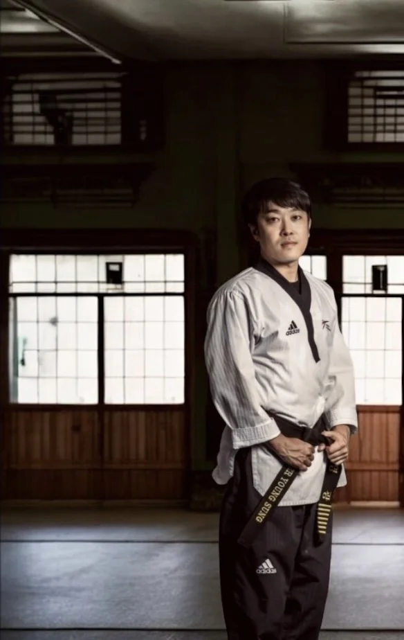 A man wearing a martial arts gi with a black belt inside a dojo with shoji-style windows.