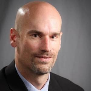 A bald man with a goatee and light skin, wearing a dark suit jacket and light blue shirt, posing against a gray background.