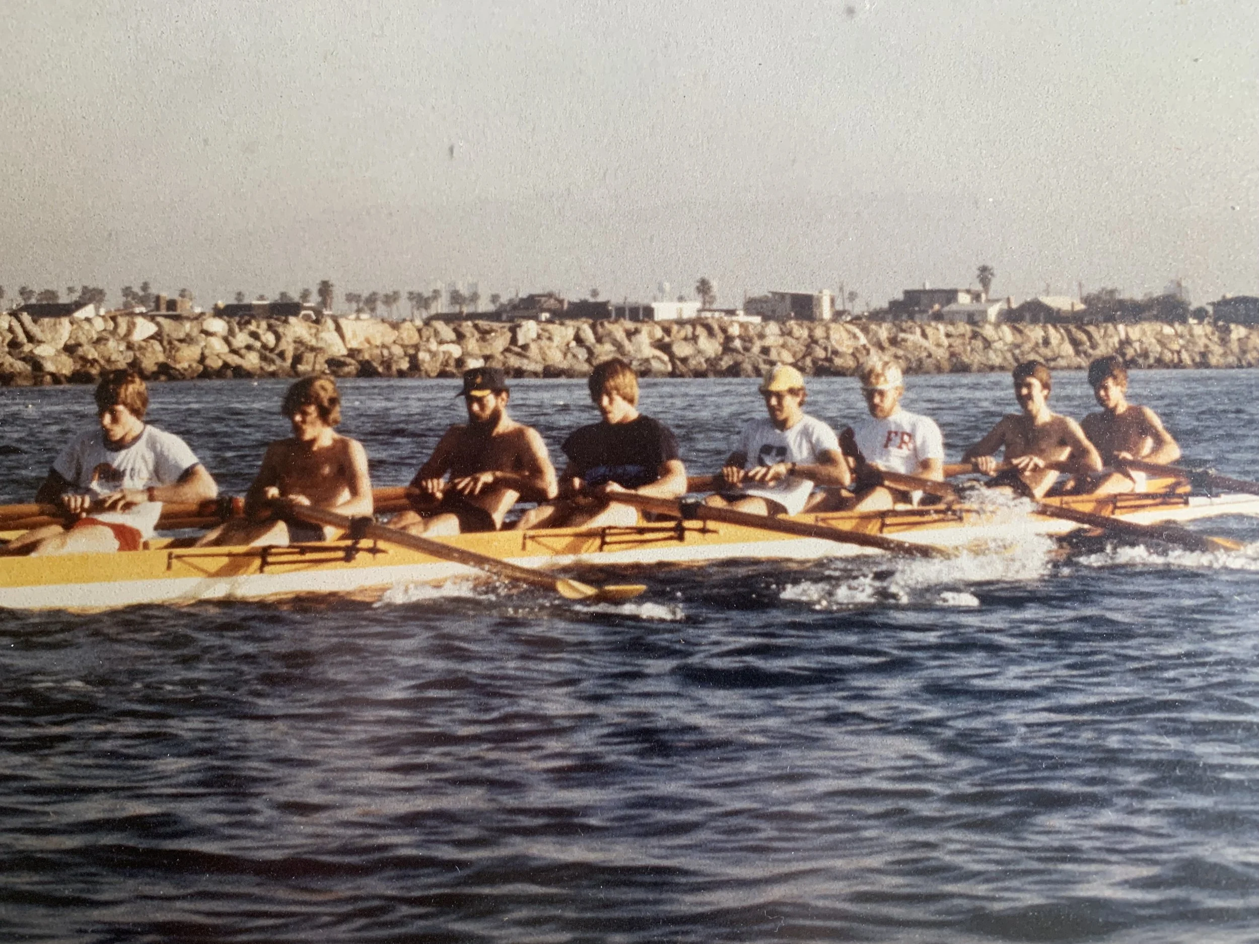 Seven young men rowing a boat on the water with houses and palm trees in the background.