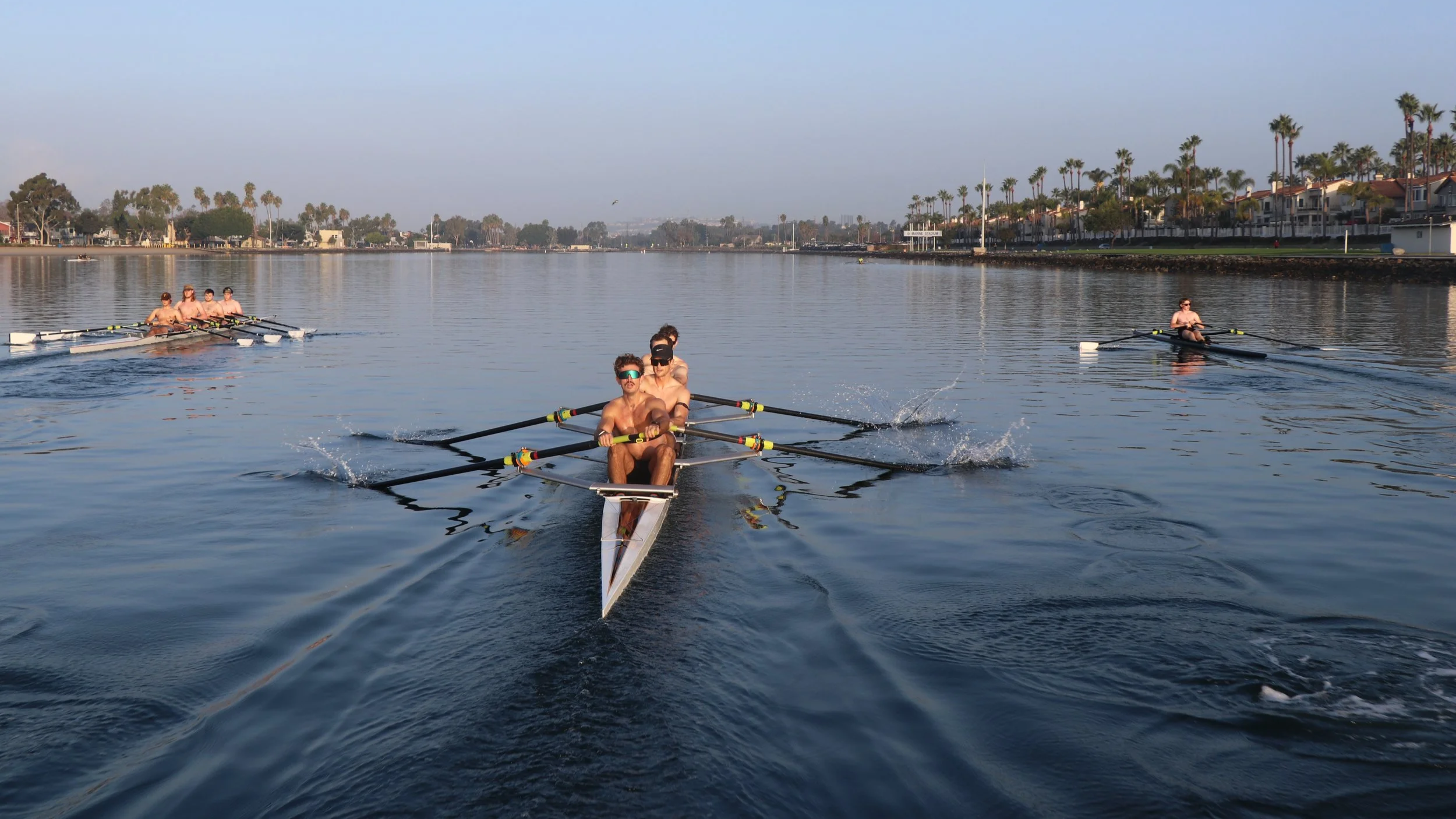 Four rowers with no shirts, wearing sunglasses, practicing rowing in a boat on a calm waterway with houses and palm trees in the background.