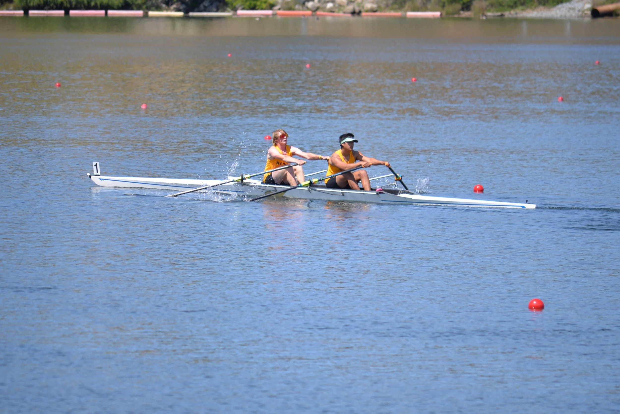 Two women in a rowing boat on a calm body of water, wearing yellow tank tops and black shorts, with some red buoys floating nearby.