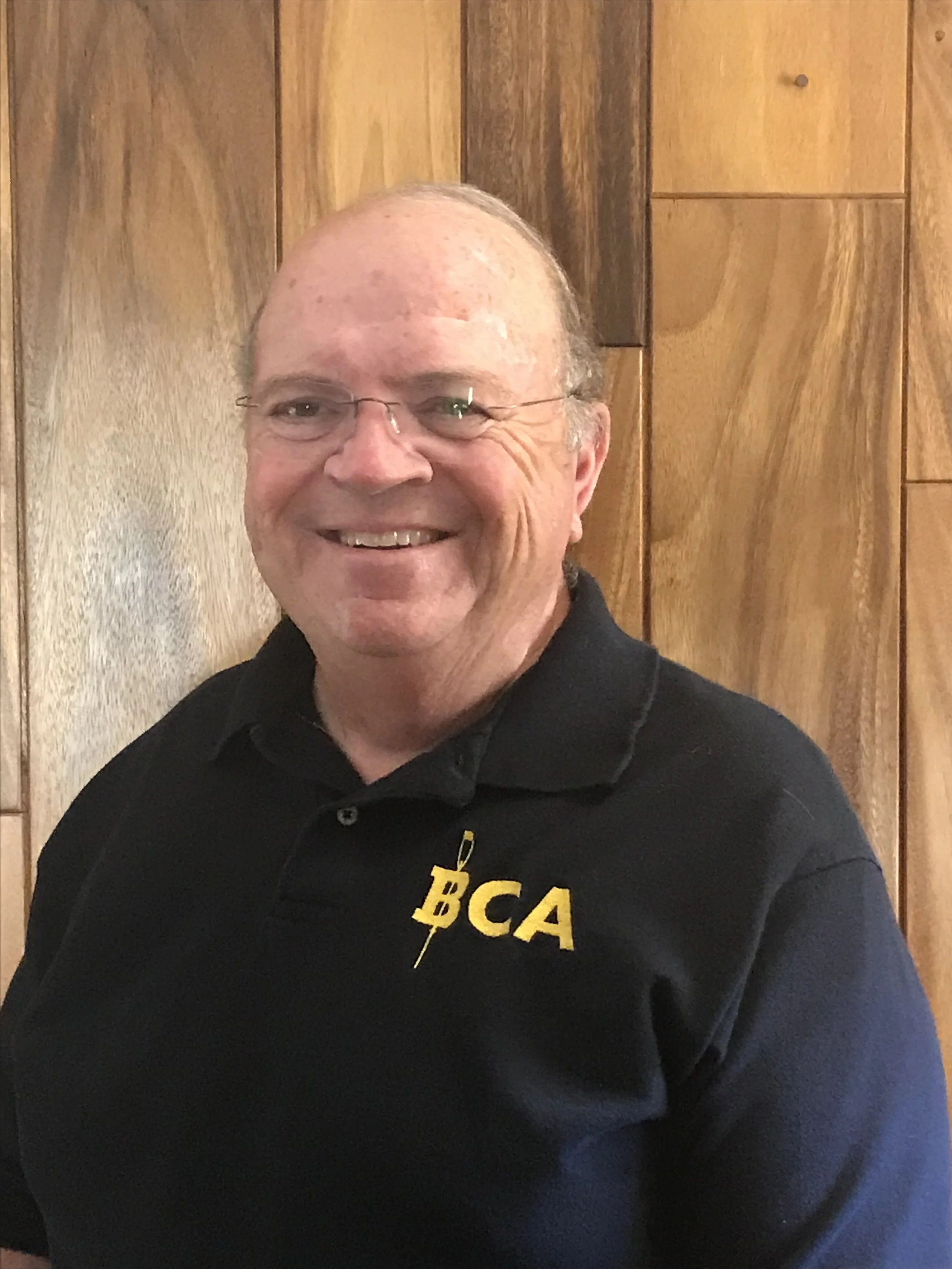 A smiling middle-aged man with glasses and receding hair, wearing a black polo shirt with yellow letters 'BCA' and a lightning bolt symbol, standing in front of a wooden wall.