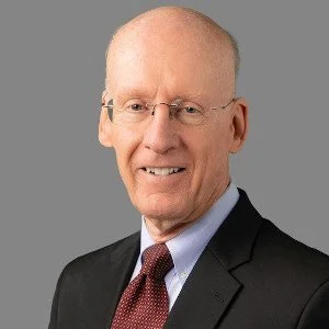 A middle-aged man with glasses, wearing a black suit, white shirt, and red tie, smiling at the camera against a gray background.