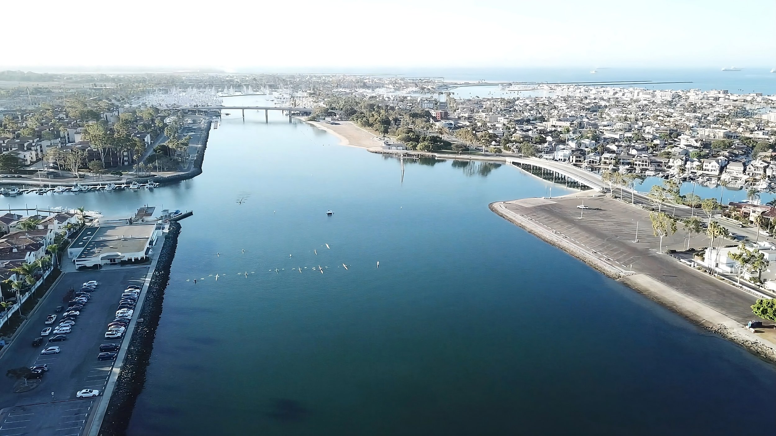 Aerial view of a coastal city with a large body of water, boats, and a marina, surrounded by residential and commercial buildings, with a glimpse of the ocean in the background.