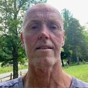 Close-up of a middle-aged man with a bald head, light beard, and wearing a gray shirt, outdoors with trees and grass in the background.