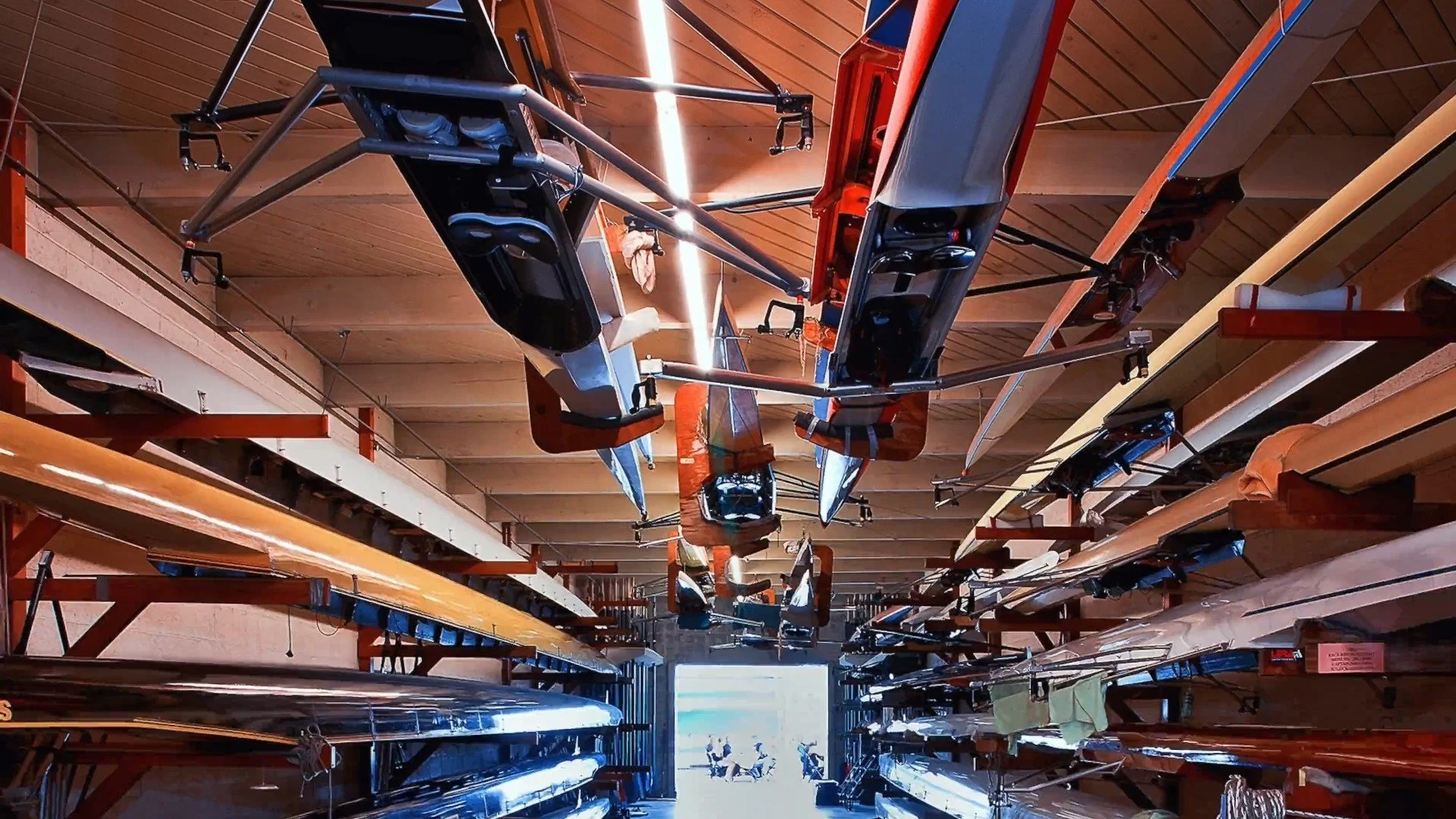 Indoor storage of racing boats on shelves with a large open door at the end showing people on jet skis outside.