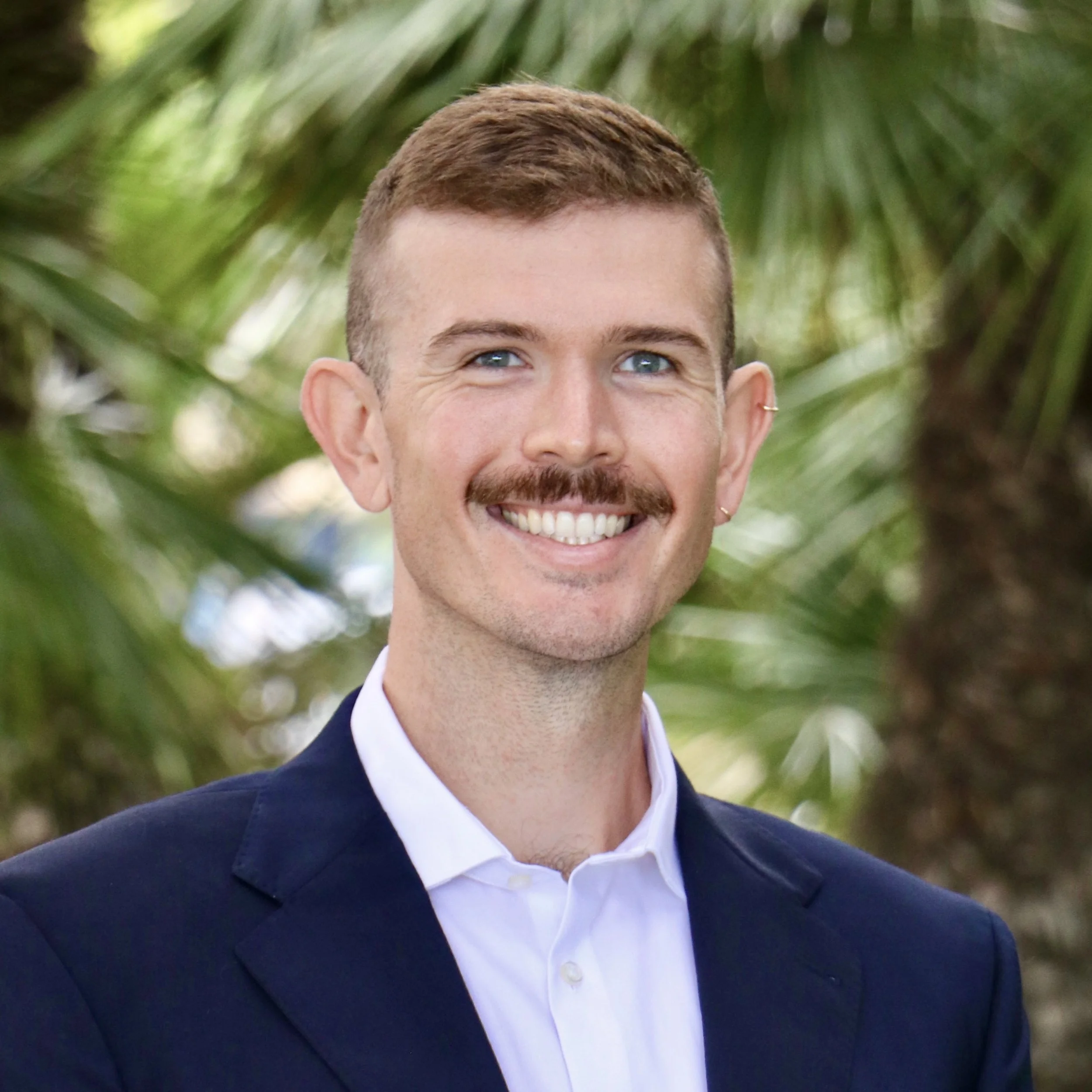 A young man with a mustache and curly brown hair, wearing a gray suit and pink tie, standing outdoors among green trees.