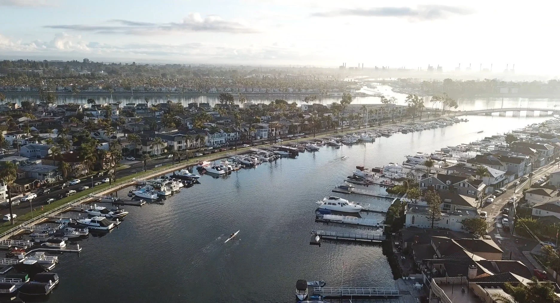 Aerial view of a marina with boats docked, residential houses, and a city skyline in the distance during daylight.