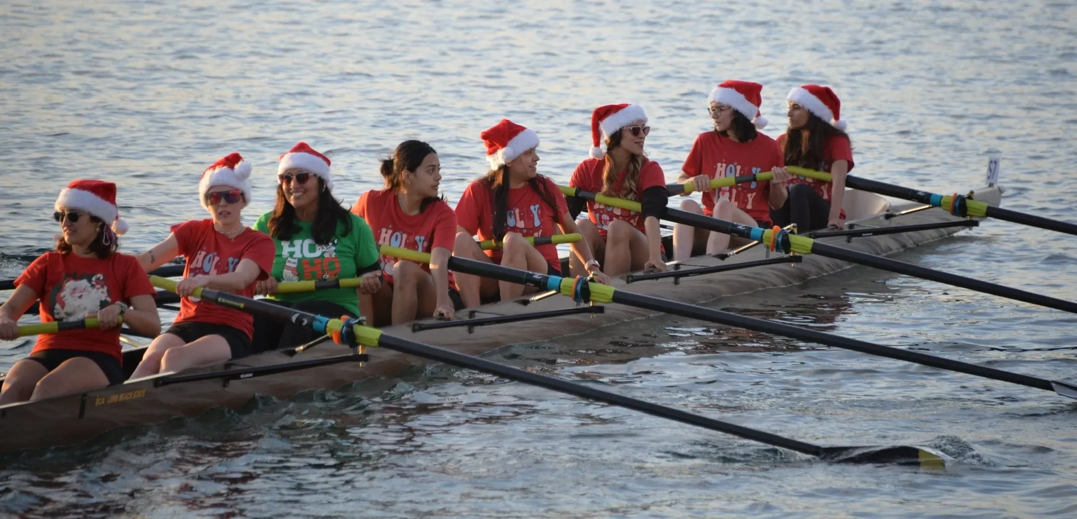 A group of women wearing Santa hats and Christmas-themed shirts, rowing a boat on the water during daytime.
