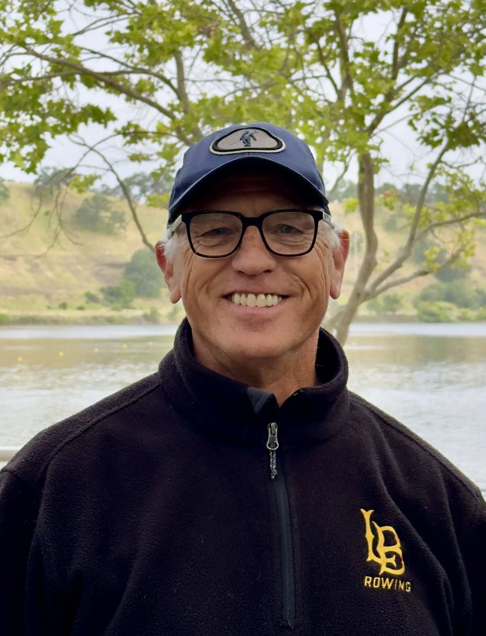 A man wearing glasses, a black cap, and a black jacket standing outdoors by a body of water with trees in the background.