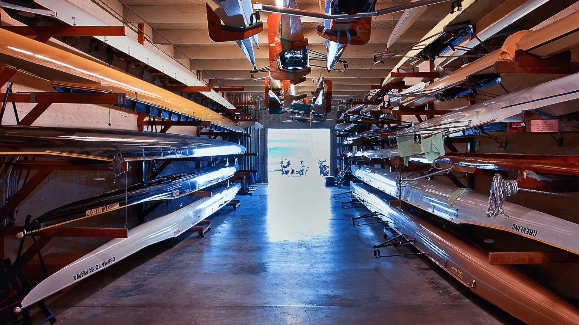 Row of racing shells and rowing boats stored on racks inside a boat storage facility, with sunlight shining through an open door at the end of the aisle. There is a small group of cyclists outside in the distance.
