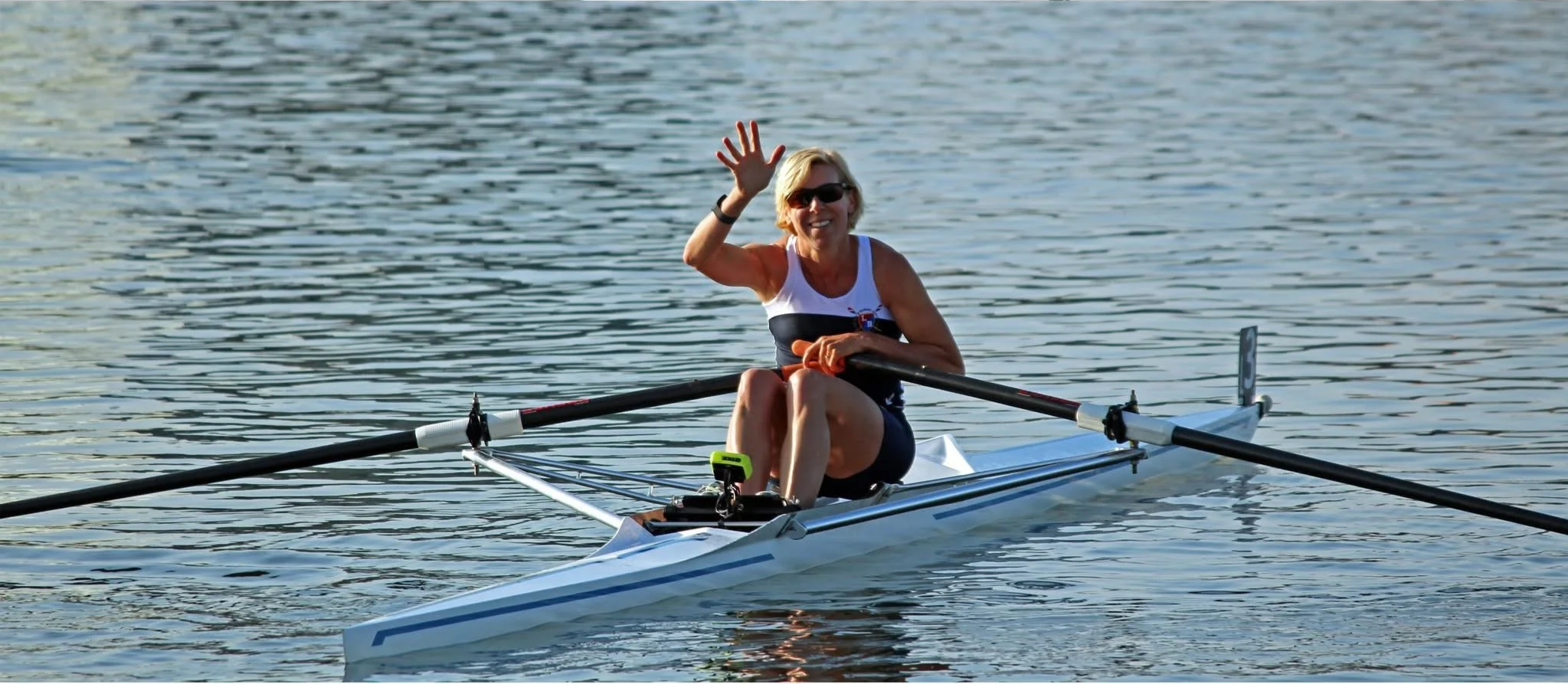 A woman in sunglasses smiling and waving while sitting in a racing rowing boat on a body of water.