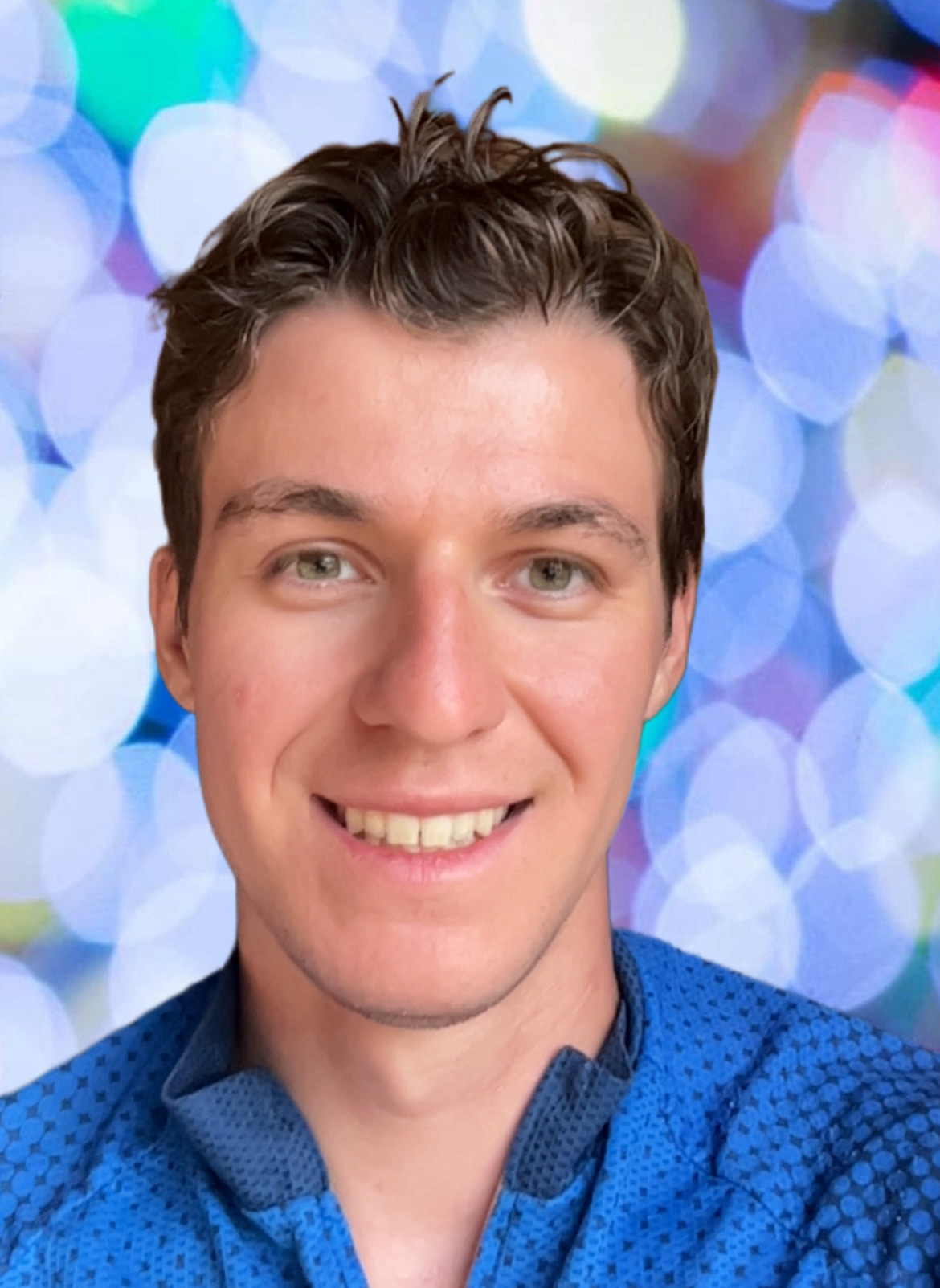A young man with brown hair and green eyes smiling, wearing a blue shirt, with a colorful, blurry background.