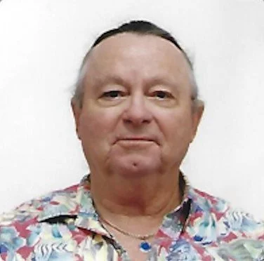 A middle-aged man with short gray hair and fair skin wearing a colorful, floral-patterned shirt, standing against a plain white background.