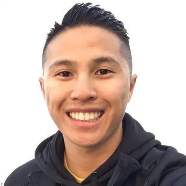 Close-up of a young man smiling, wearing a black hoodie and standing against a plain white background.