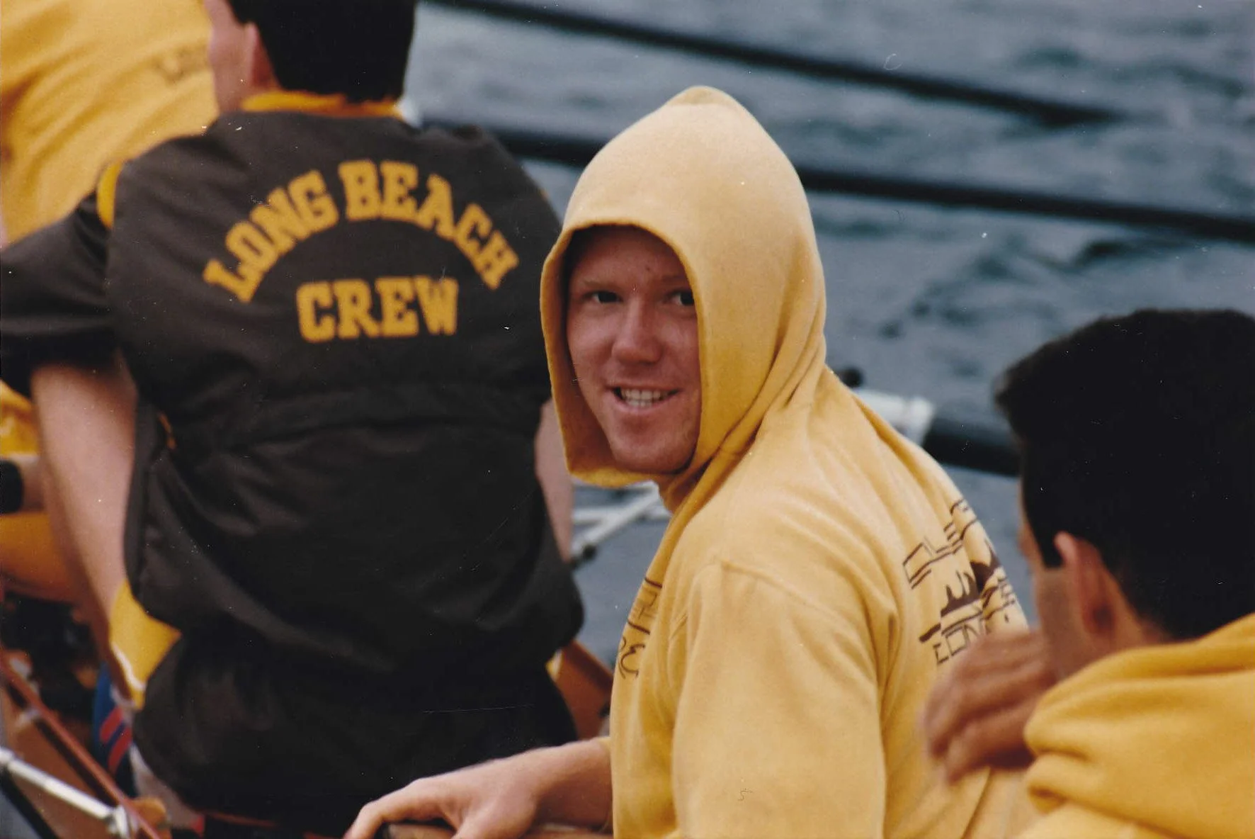 A smiling man wearing a yellow hoodie during a boat race, with the water in the background and other crew members around him.