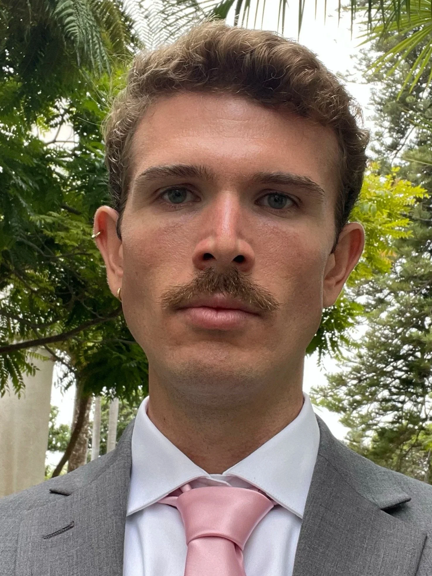 A young man with a mustache and curly brown hair, wearing a gray suit and pink tie, standing outdoors among green trees.