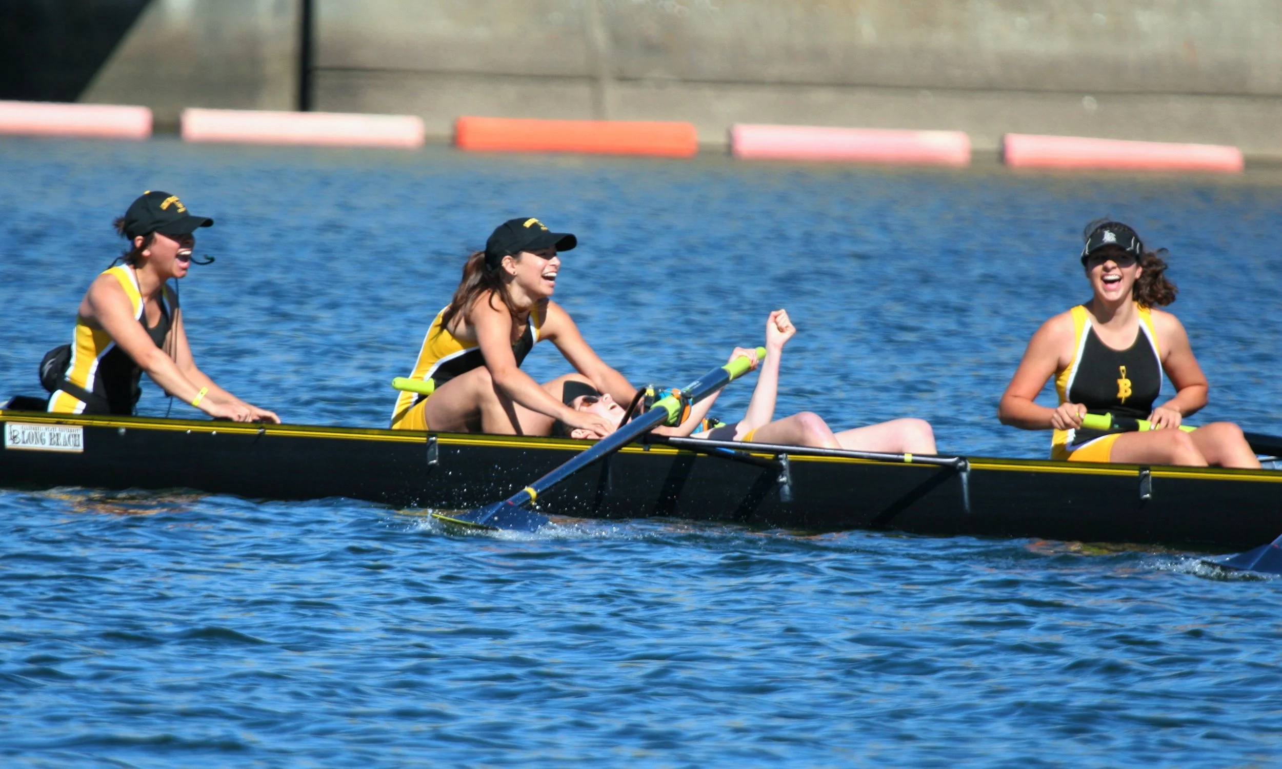 Women in yellow and black athletic uniforms participating in a rowing competition on a blue water body. They are smiling and celebrating, with one woman lying on the boat, raising a fist. The spectators or dock are visible in the background.