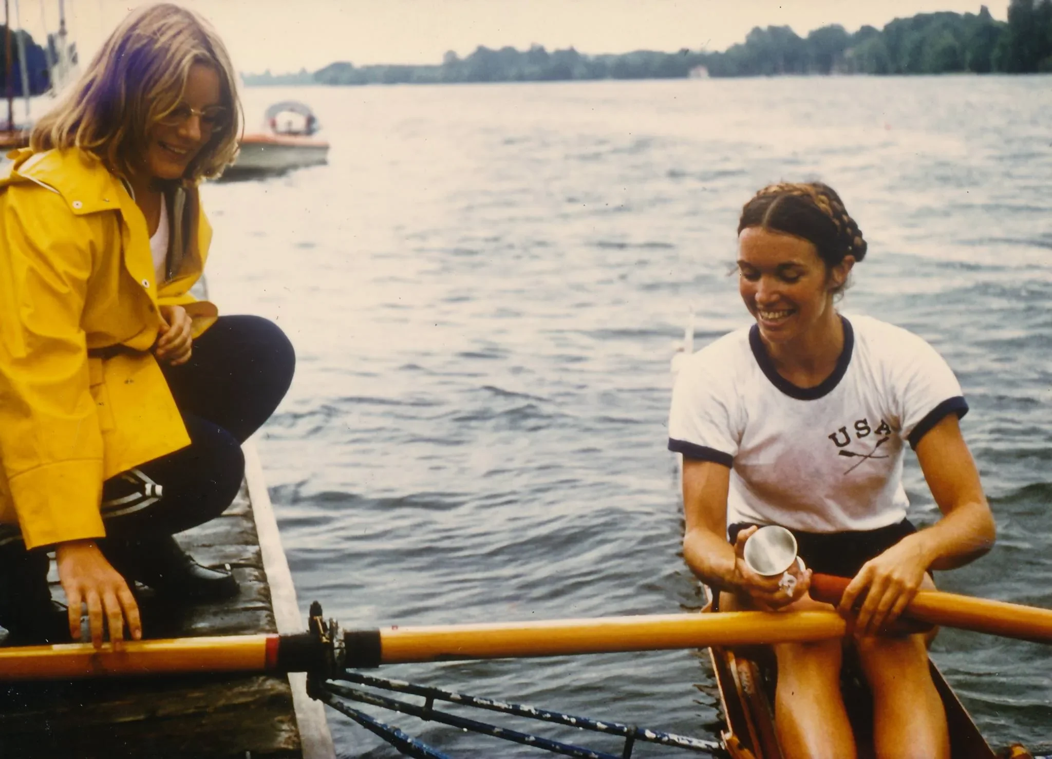 Two women on a dock by a river, one sitting in a boat with a paddle and the other crouching on the dock, both smiling. The woman in the boat is wearing a white USA t-shirt with black trim, and the woman on the dock is wearing a yellow rain jacket.